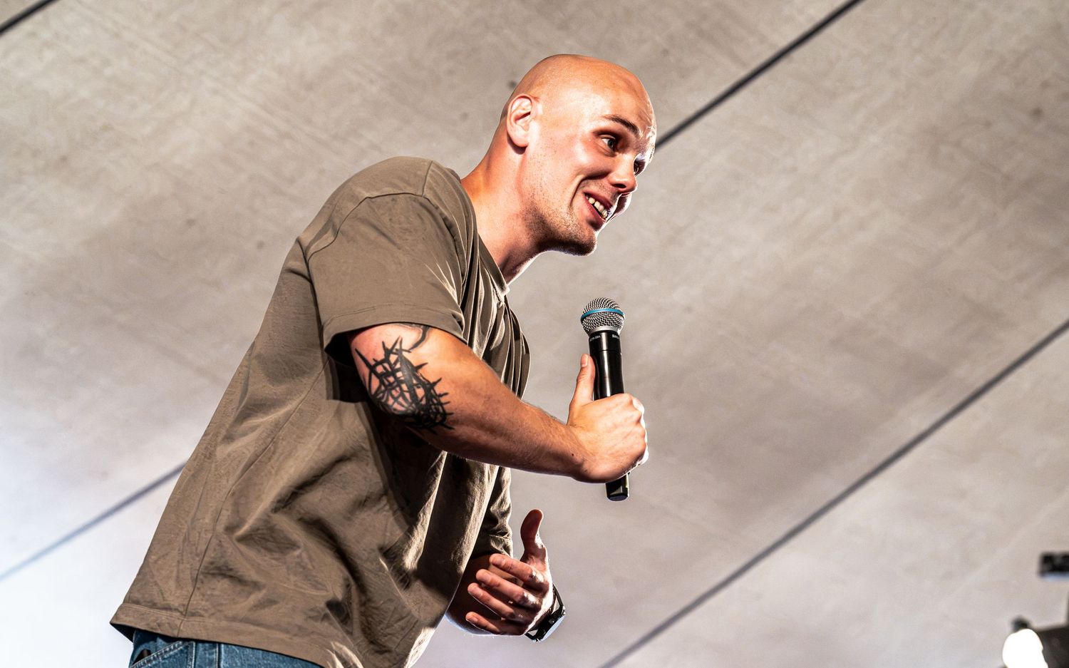 Tattooed bald male performer singing passionately into a microphone on stage wearing an olive green t-shirt.