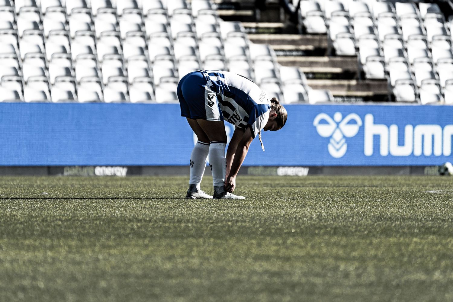 Female soccer player in dark kit bending down to tie shoelaces on an empty stadium pitch near Hummel ad.