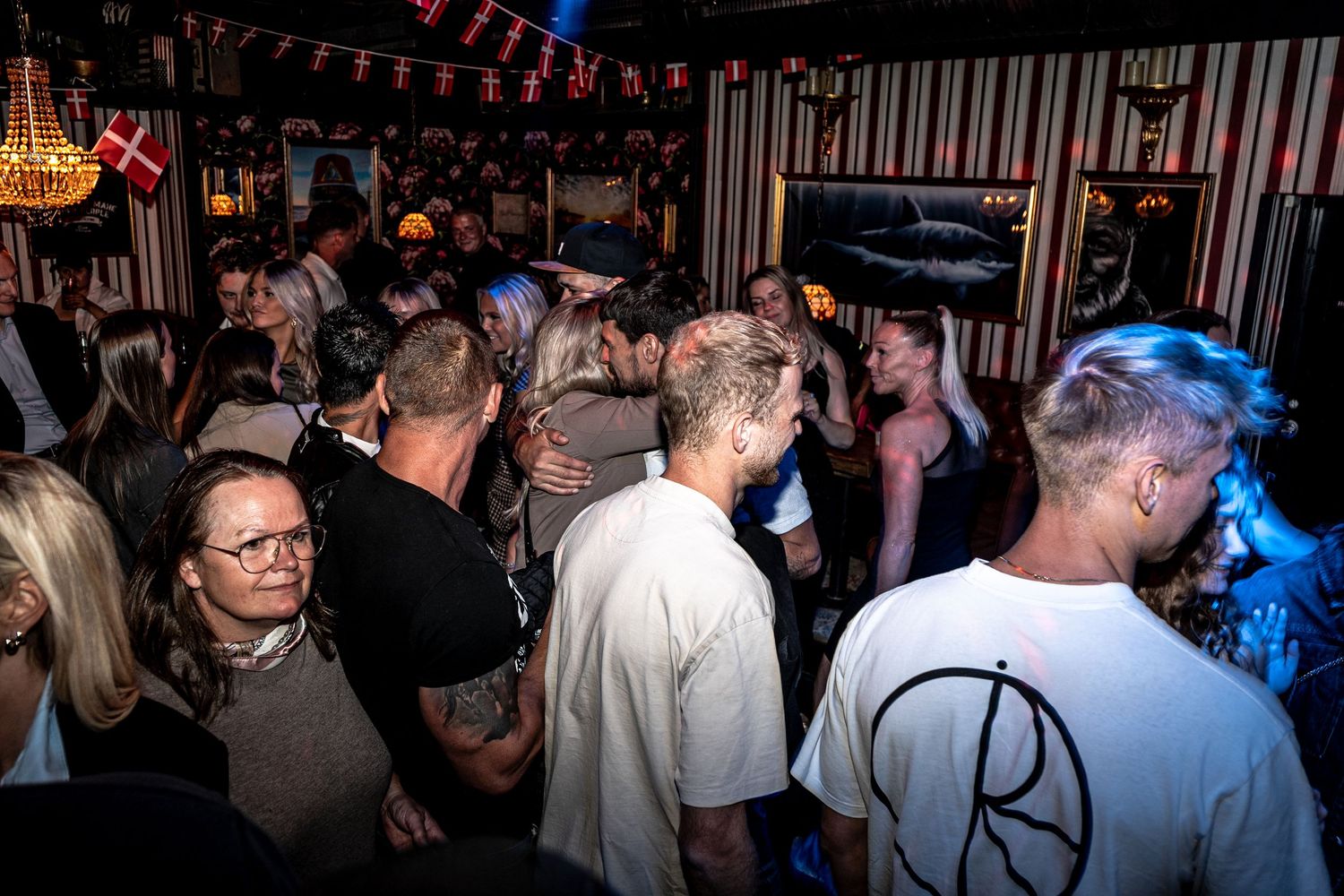 Crowded nightlife scene with people socializing in a dimly lit bar with striped walls, a chandelier and a Danish flag in the background.