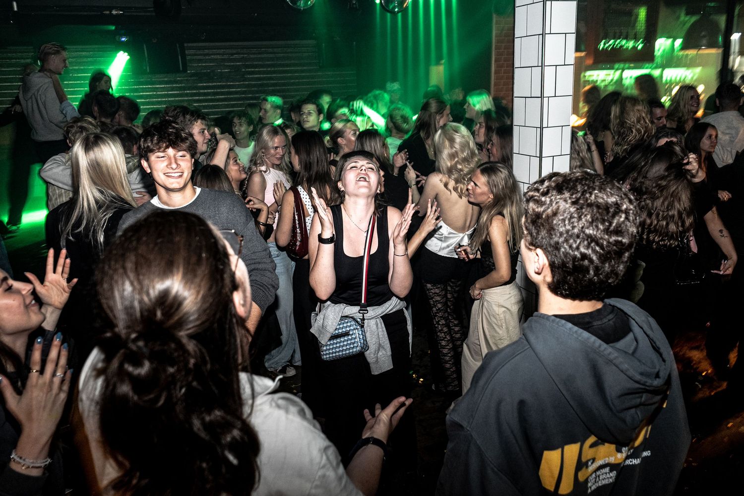 A lively crowd dances and cheers in a nightclub bathed in green light, with a young man smiling in the center.