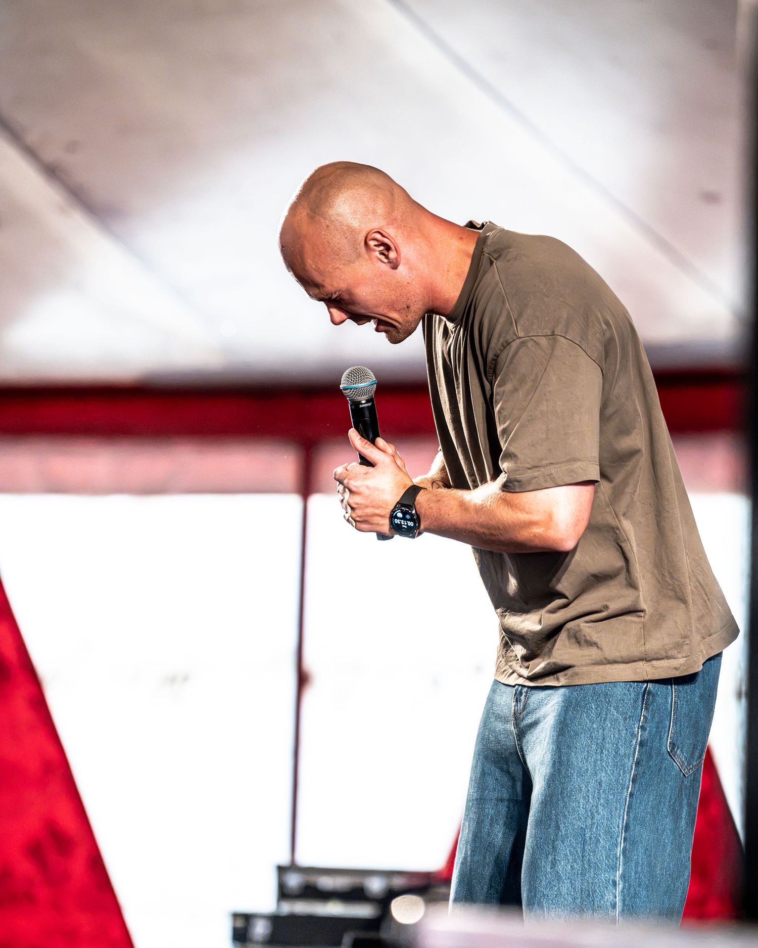 Bald male performer bends forward holding microphone on stage at outdoor festival with red and white tent backdrop.