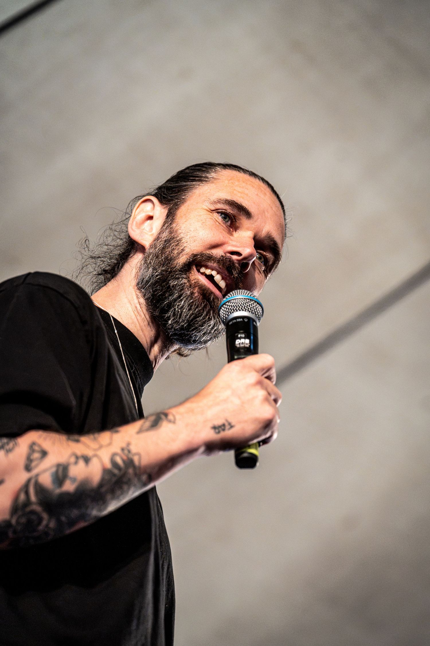 Tattooed bearded male singer performs energetically with microphone on outdoor festival stage.