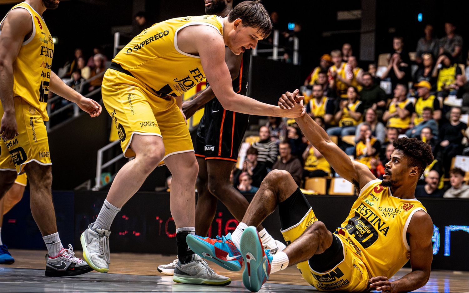 Teammate helps fallen yellow-uniformed basketball player up during an intense league game.
