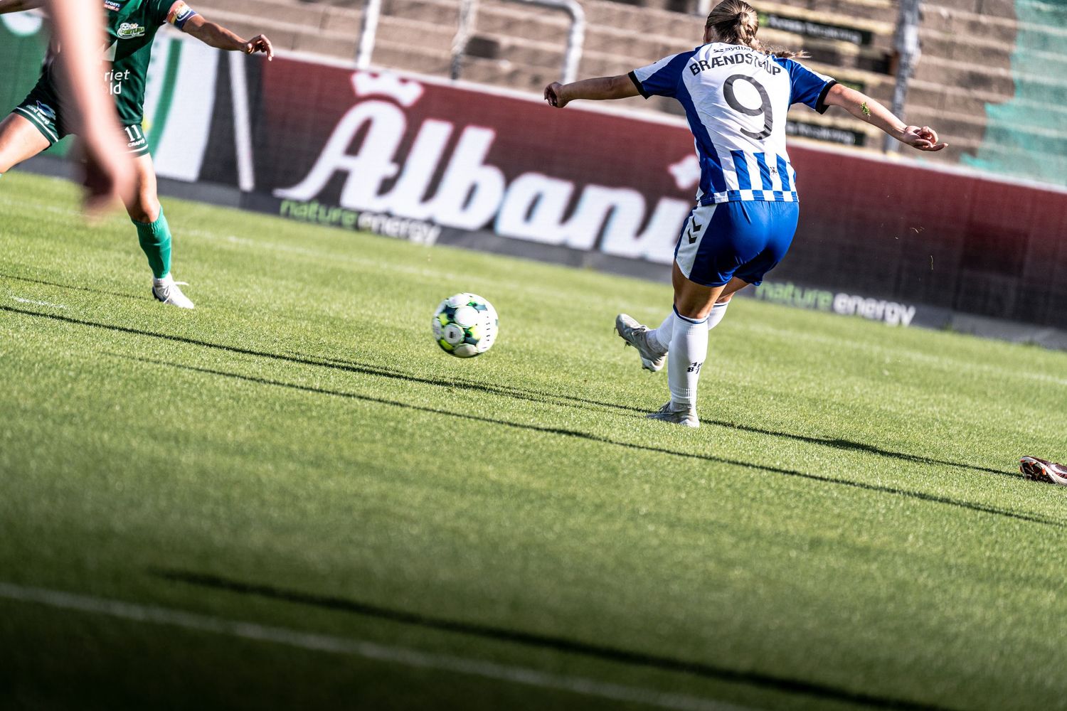 Female soccer player wearing number 9 blue and white jersey kicks ball during competitive match on green field.