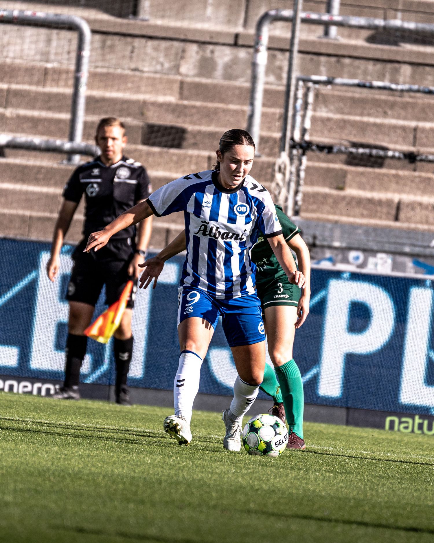 Female soccer player wearing number 9 in blue and white Alsani kit dribbling ball during a league match.