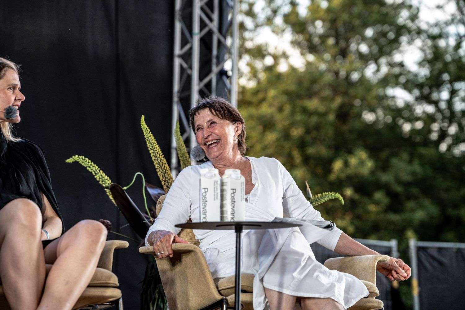 Woman in white outfit laughing joyfully while seated at an outdoor stage panel discussion.