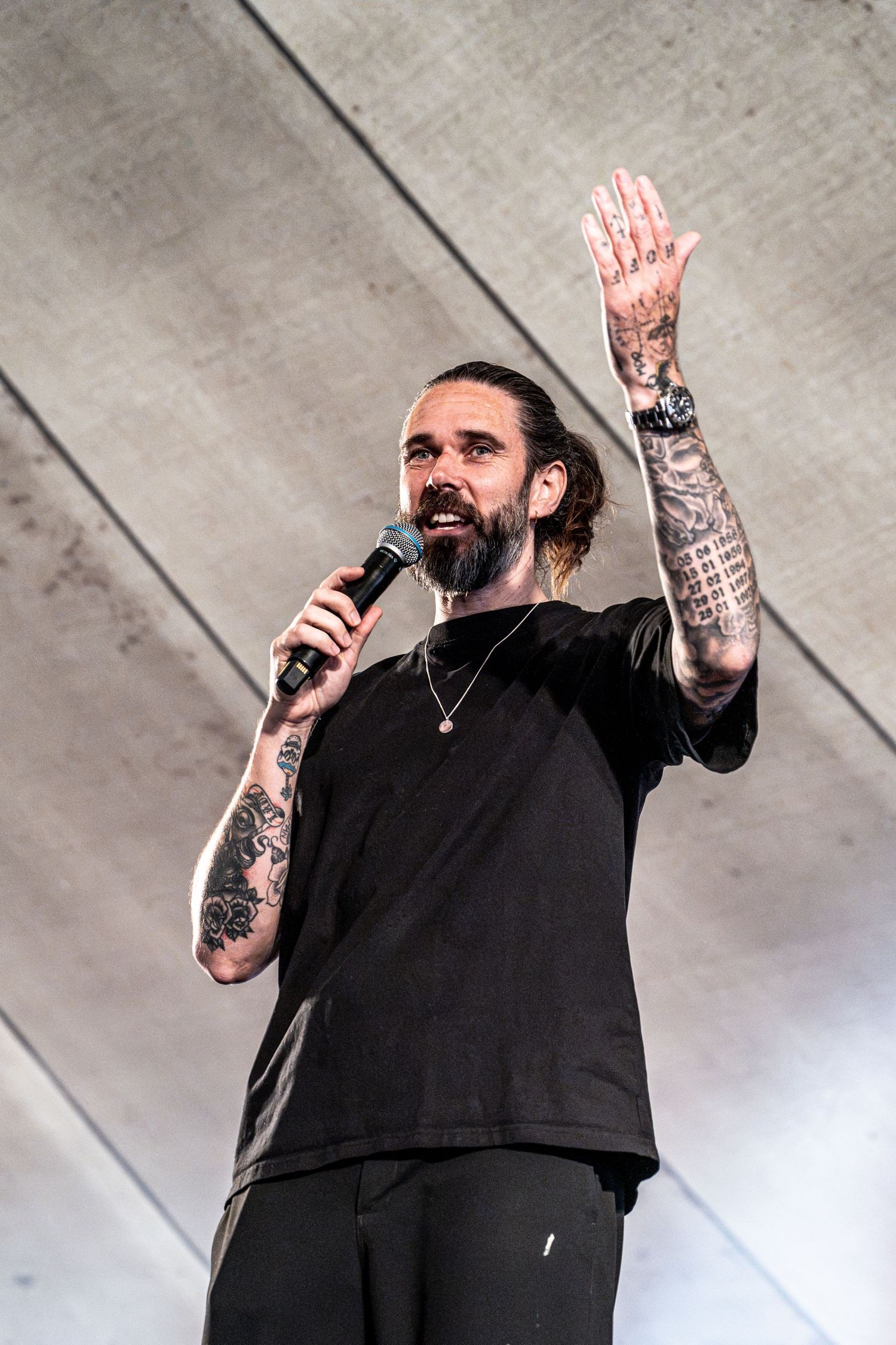 Tattooed male performer raising hand while speaking into microphone on outdoor festival stage.
