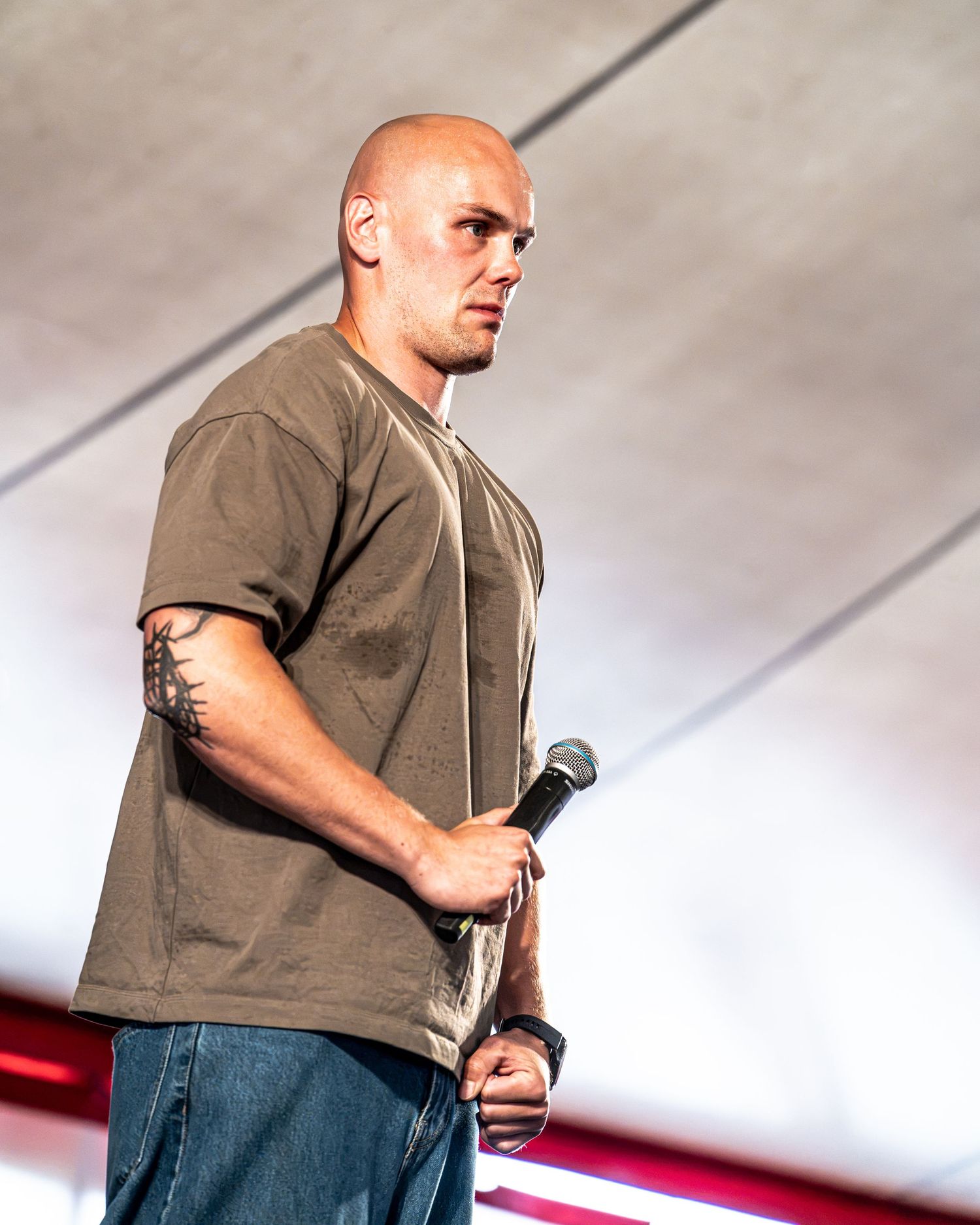 Bald man with arm tattoo standing on stage holding microphone in olive t-shirt and jeans.