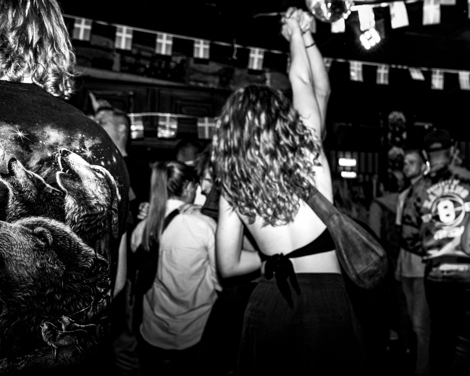 Black and white photo of a woman with curly hair dancing with raised arms in a crowded pub under string flags.