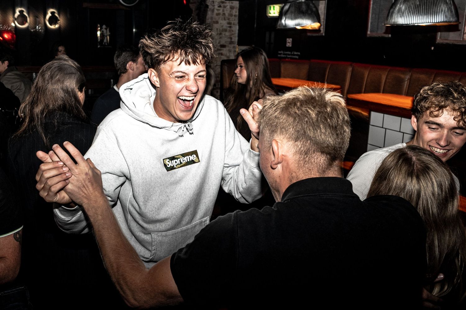 Laughing young man in a gray hoodie dances with friends in a lively, dimly lit bar while others celebrate around him.