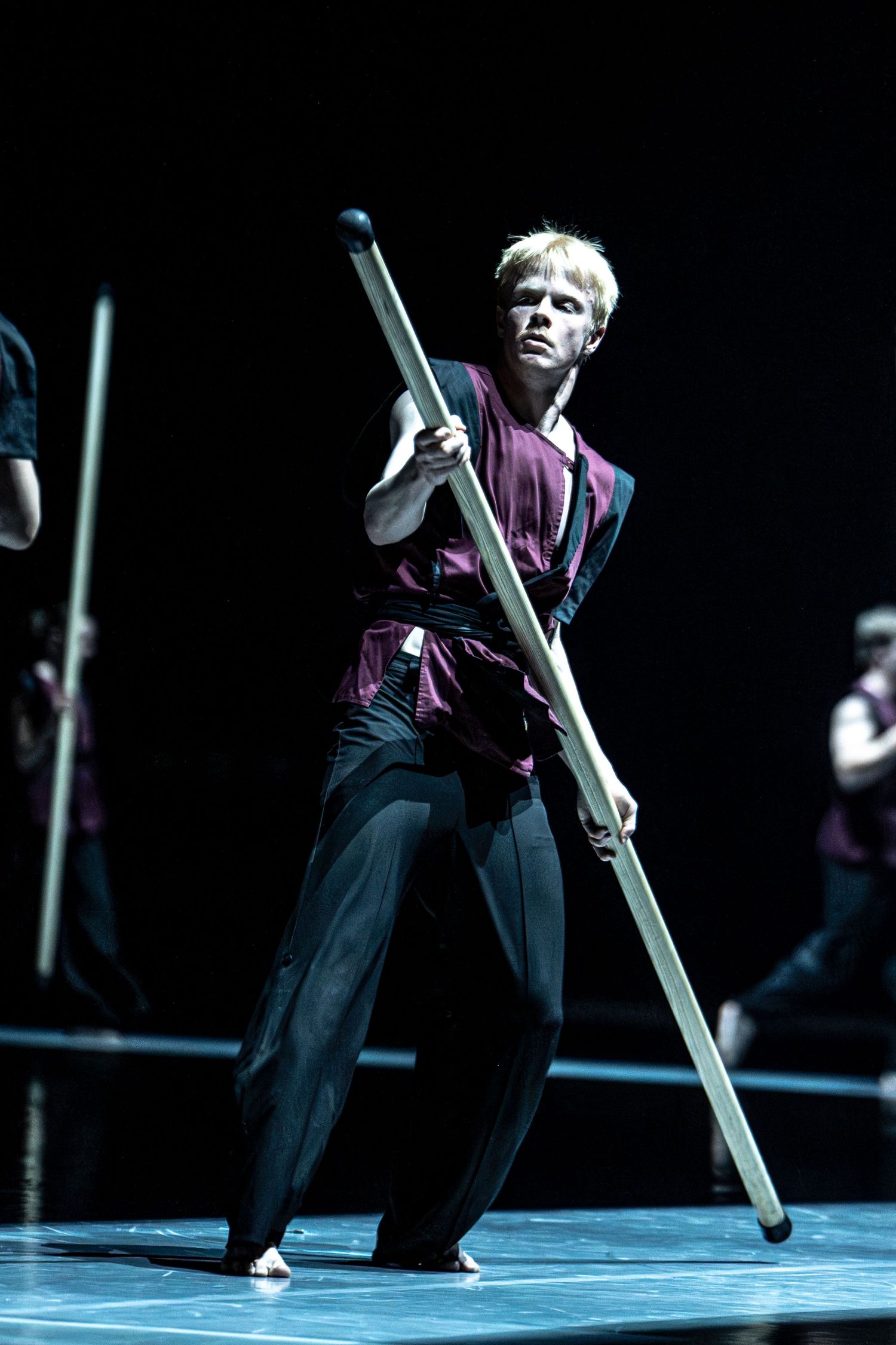 Blonde male dancer in burgundy costume performs with long staff on dramatic dark stage.