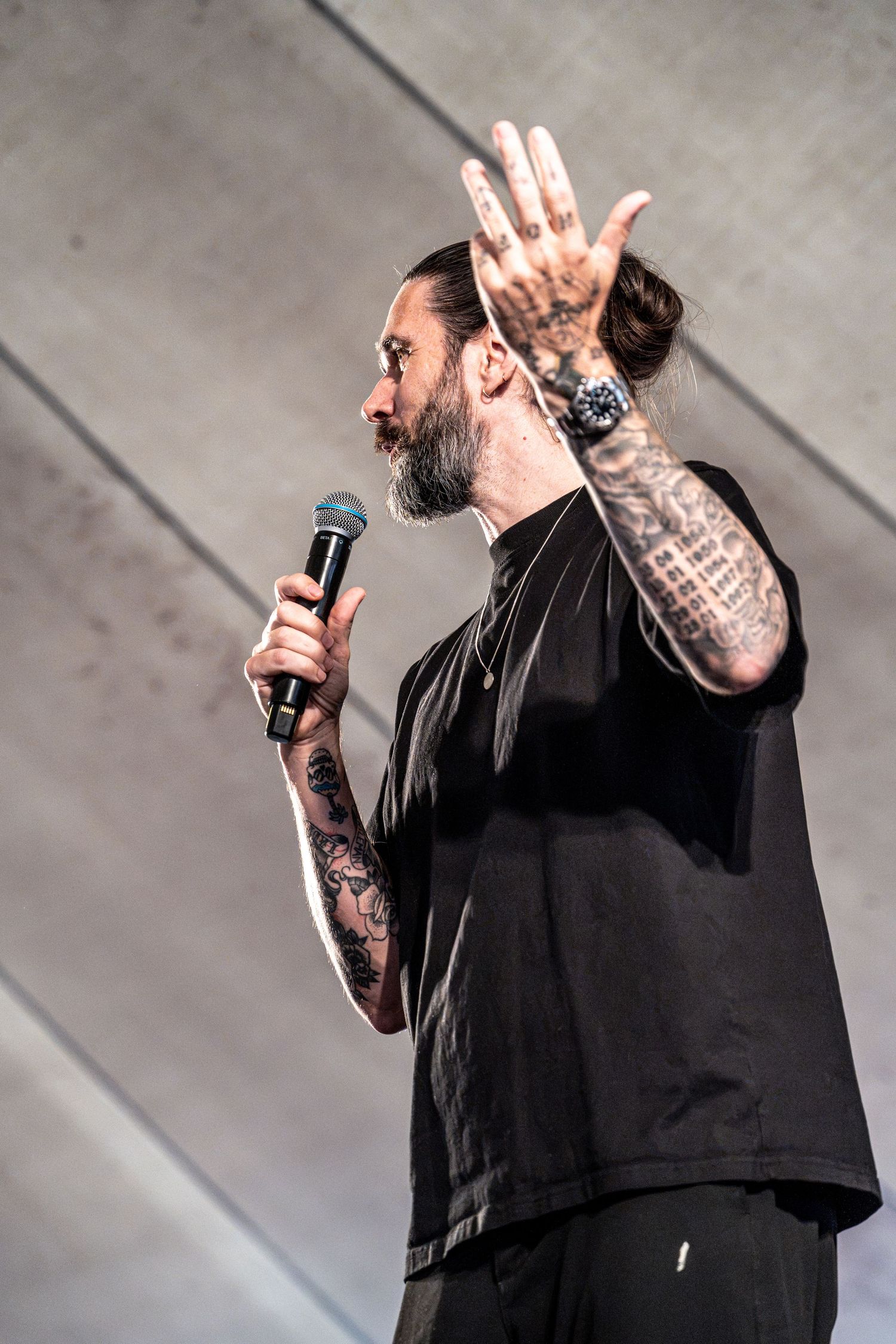 Tattooed bearded male singer raising open palm while performing on stage holding a microphone.