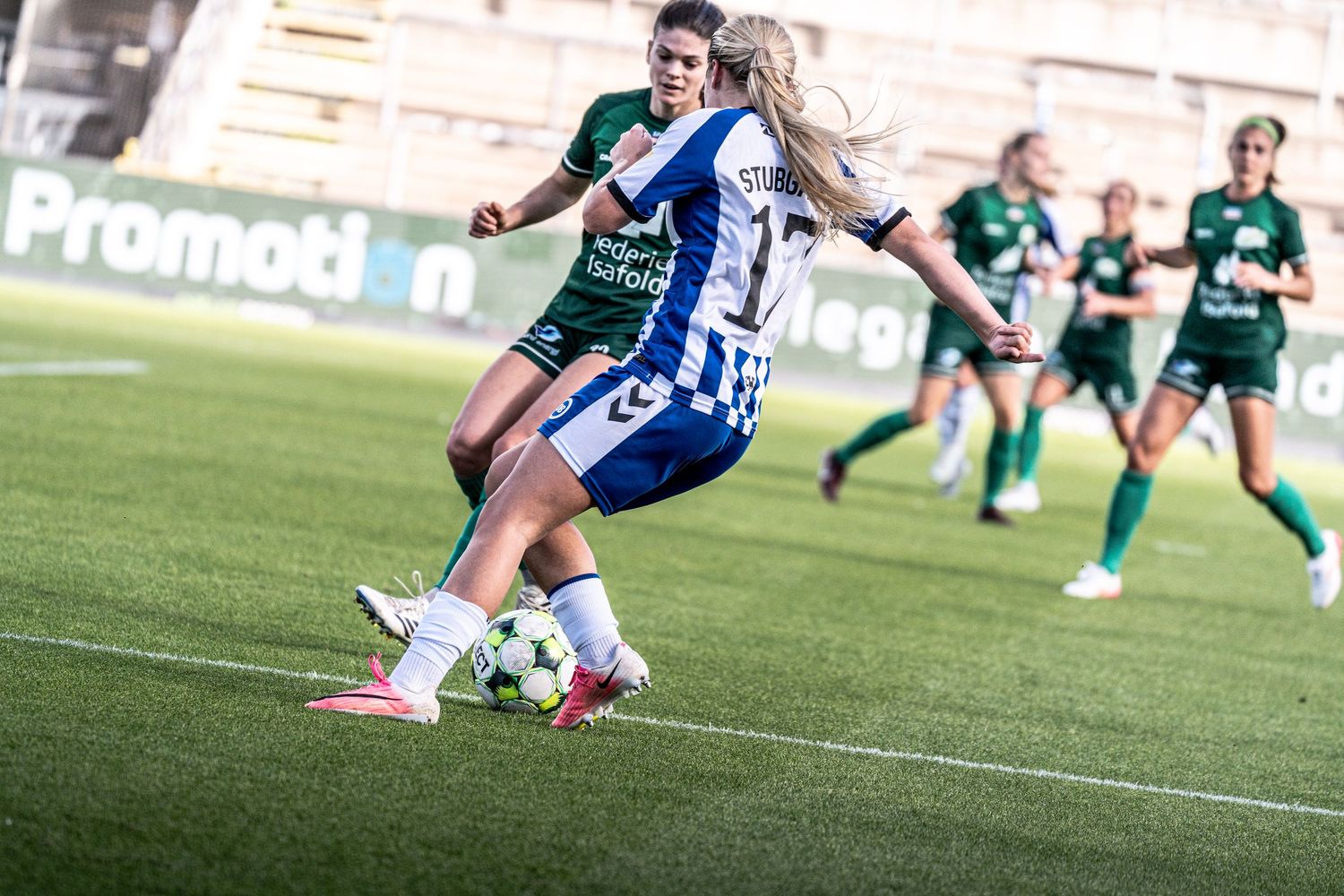 Women's soccer player #17 in blue and white shields the ball from a green-kitted defender on the pitch.