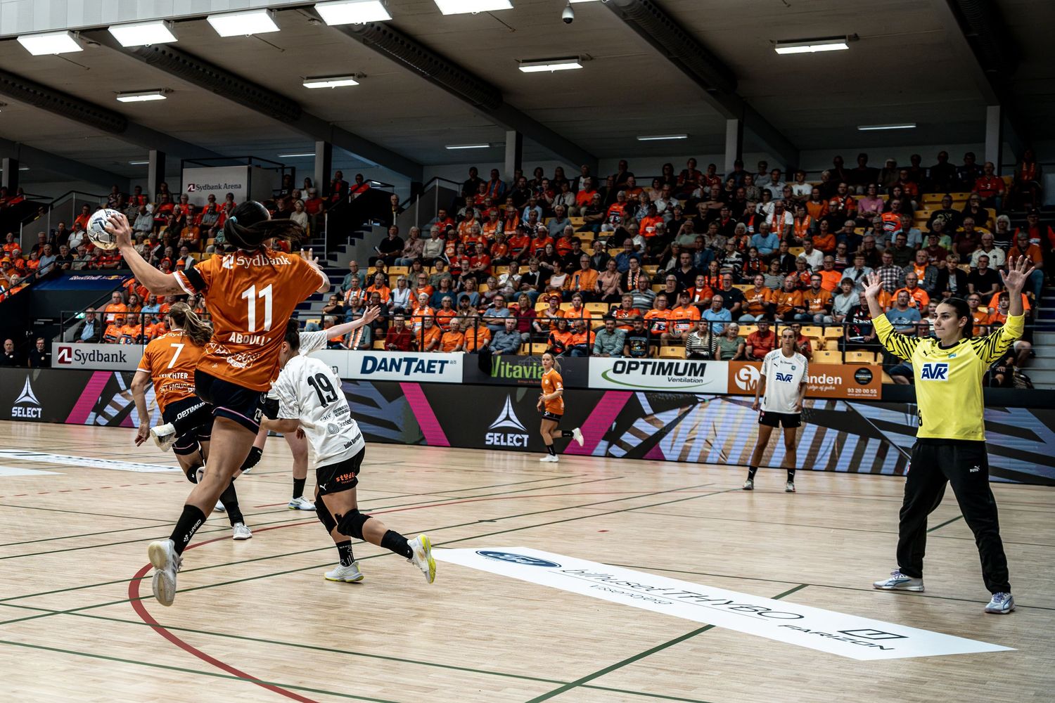 Handball player number 11 shoots at goal during an indoor handball match with orange-clad home supporters.