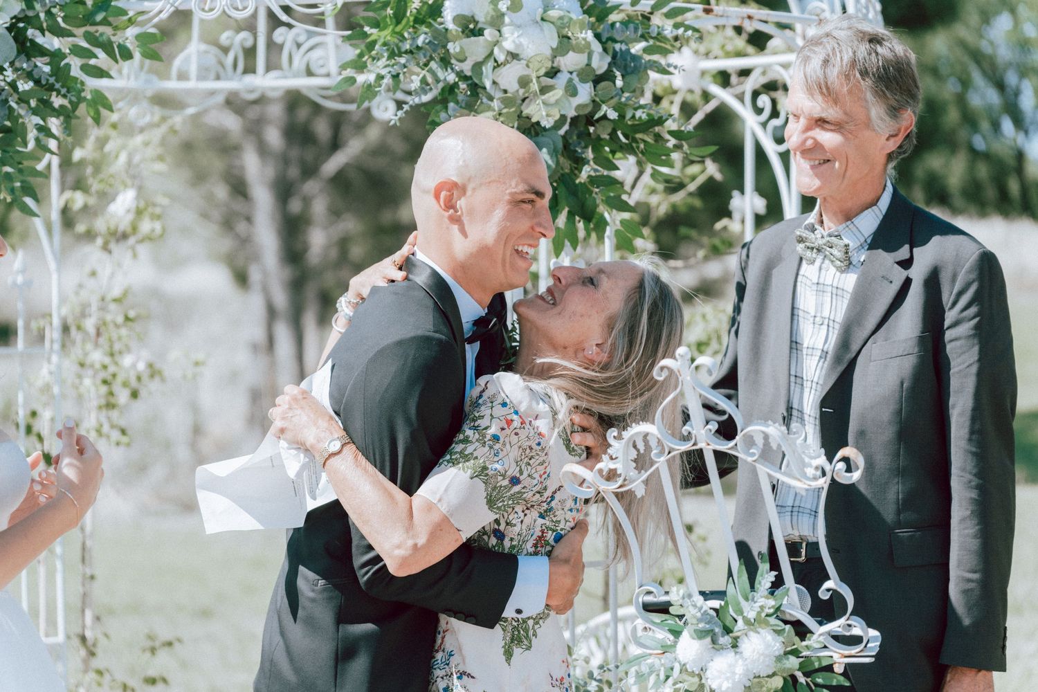 Groom embracing mother with joyful smiles beside officiant at outdoor wedding ceremony.