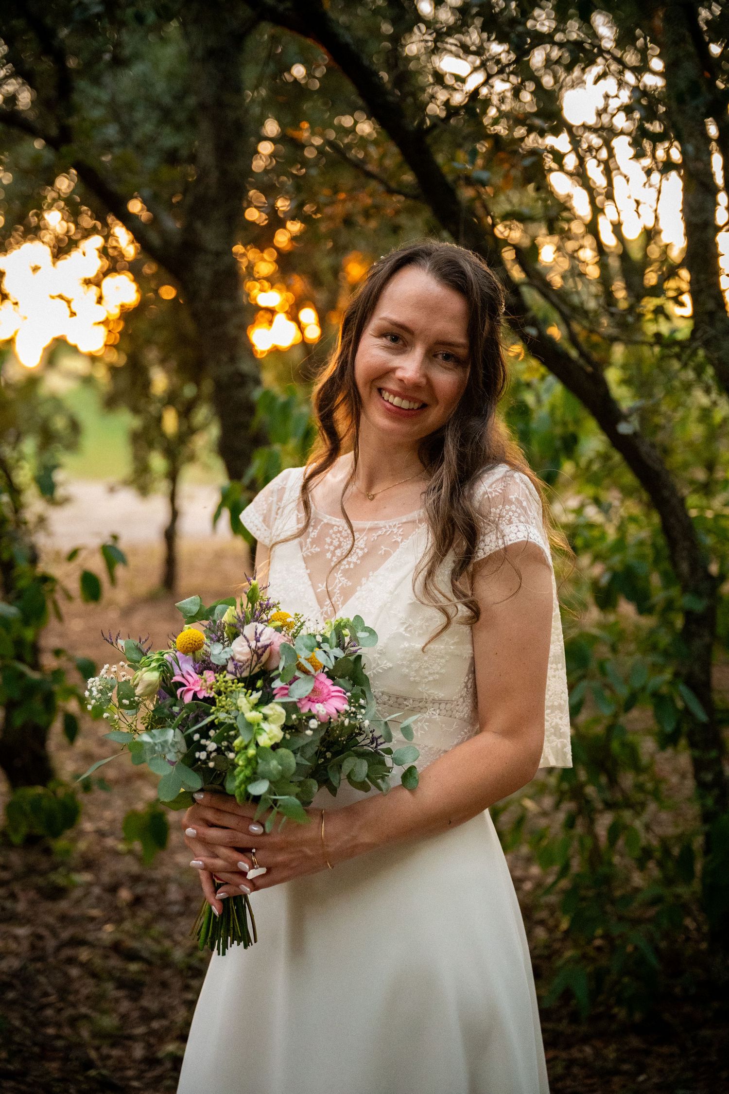 Smiling bride in lace white dress holding colorful wildflower bouquet, posing outdoors at golden hour.