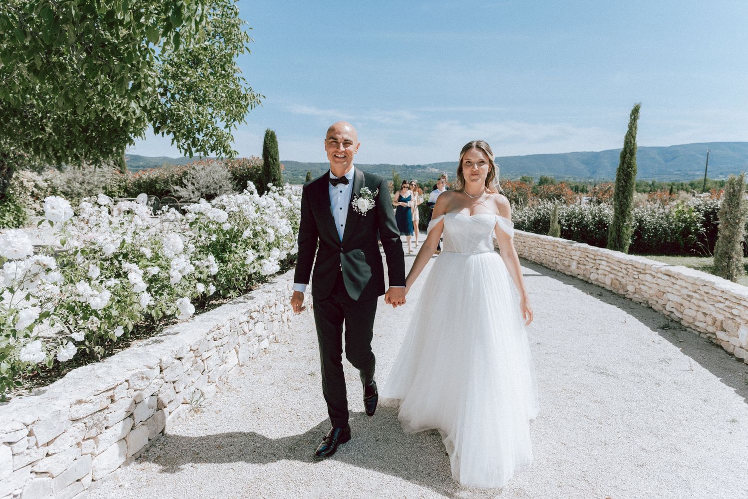 Newlyweds holding hands walking down a white stone path lined with white flowers on a sunny day.