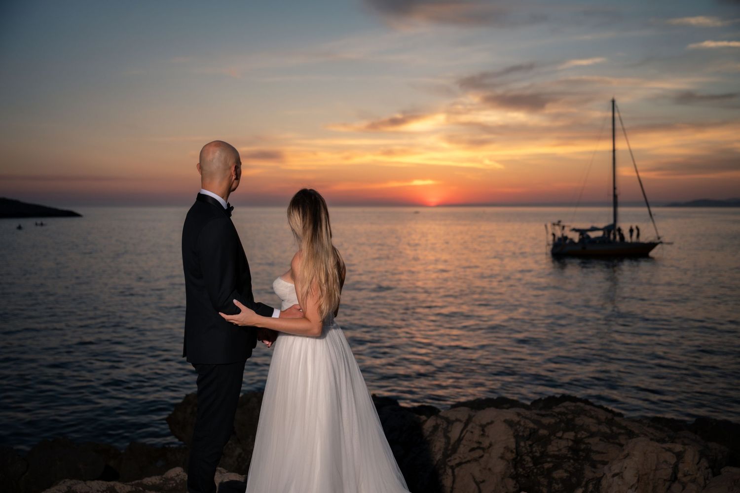 Bride and groom holding hands at rocky seaside with sailboat and vibrant sunset sky.