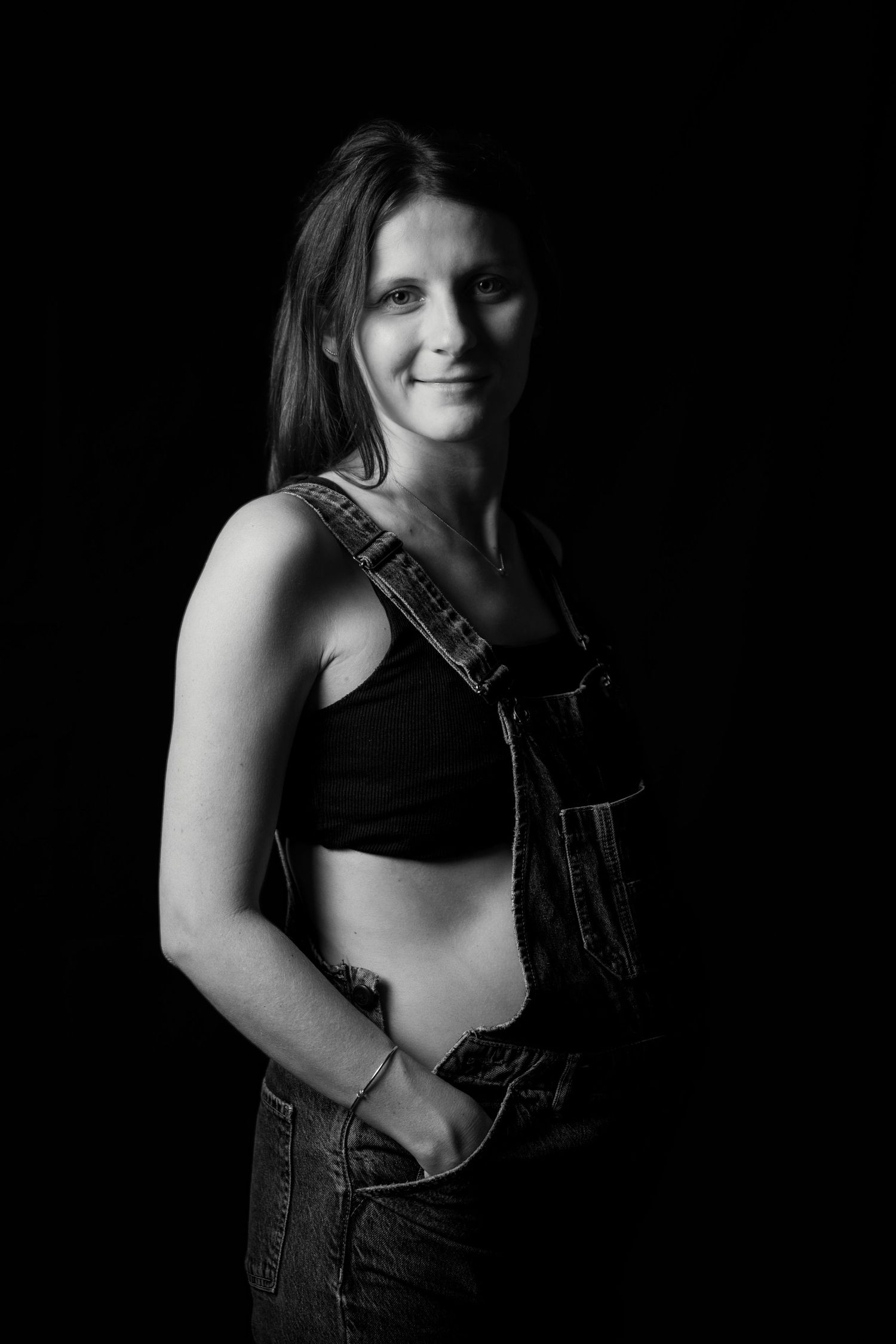 Black and white portrait of a pregnant woman in denim overalls smiling softly at the camera in a studio setting.