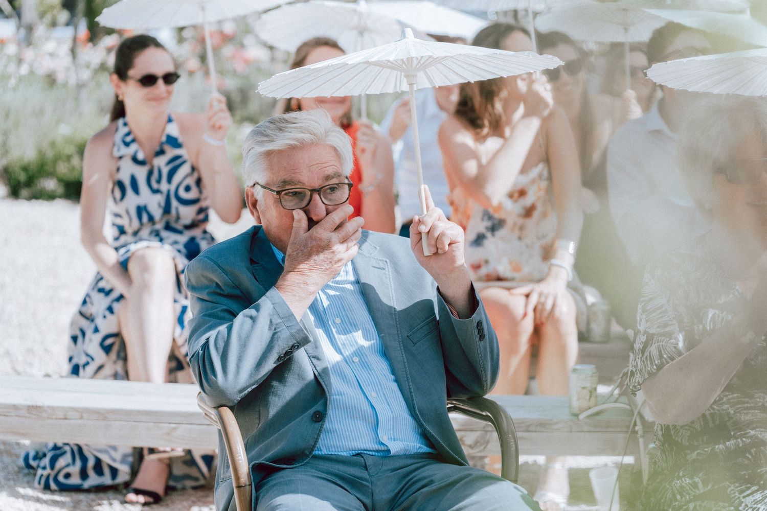 Elderly gentleman in blue suit sips a drink at an outdoor wedding reception under parasols.
