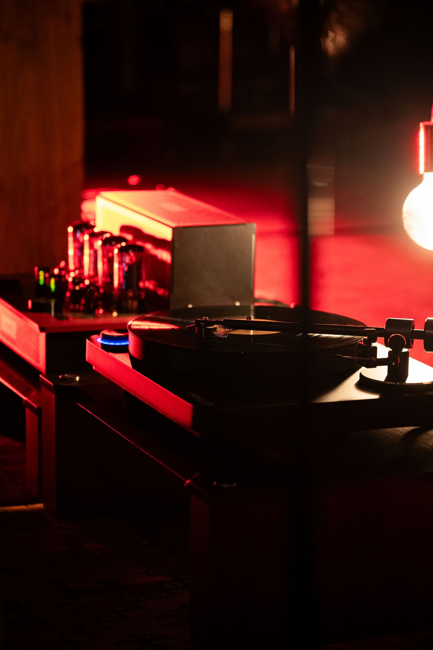Vintage turntable playing vinyl record in dimly lit room with red ambient lighting and audio equipment in background.