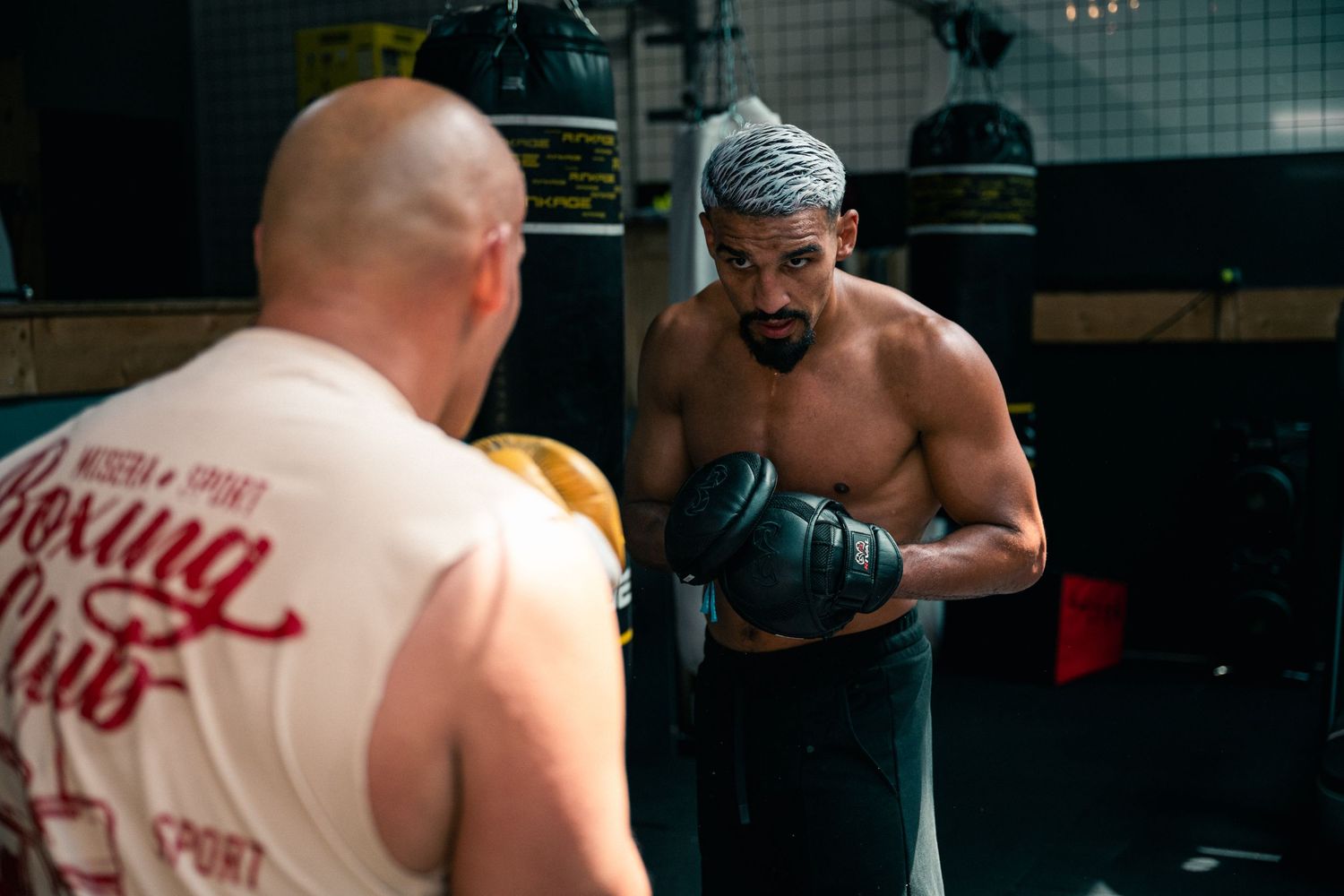 Two boxers sparring in a gym, one shirtless with gray-streaked hair wearing green gloves in fighting stance.