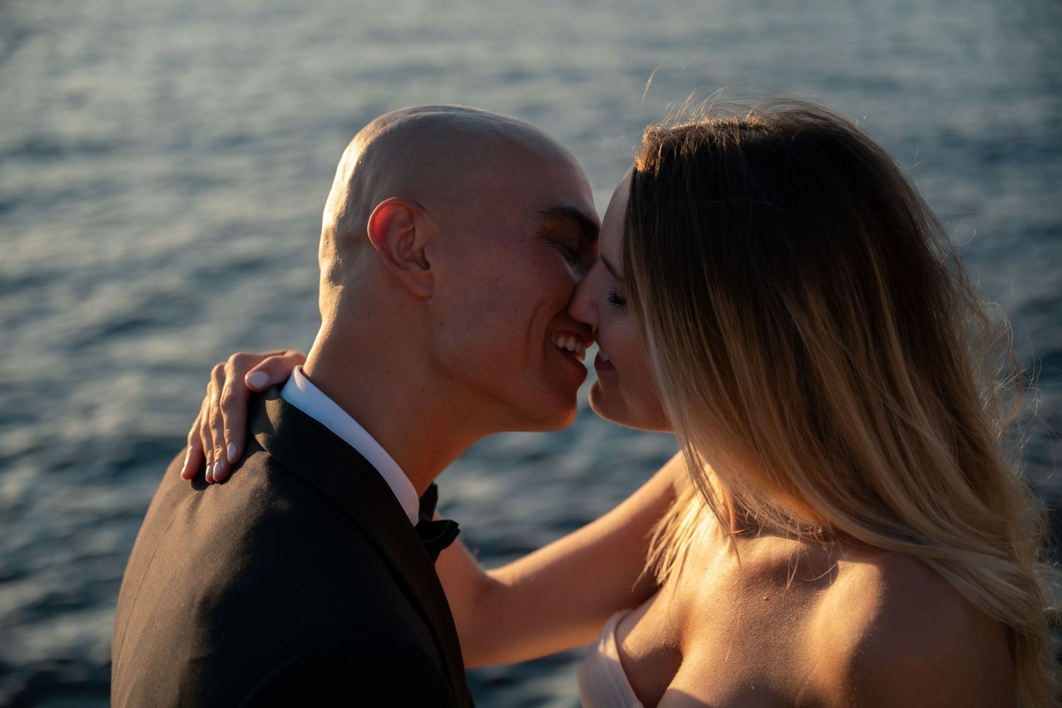Couple sharing a romantic almost-kiss near the ocean at sunset, man in tuxedo embracing woman in white dress.