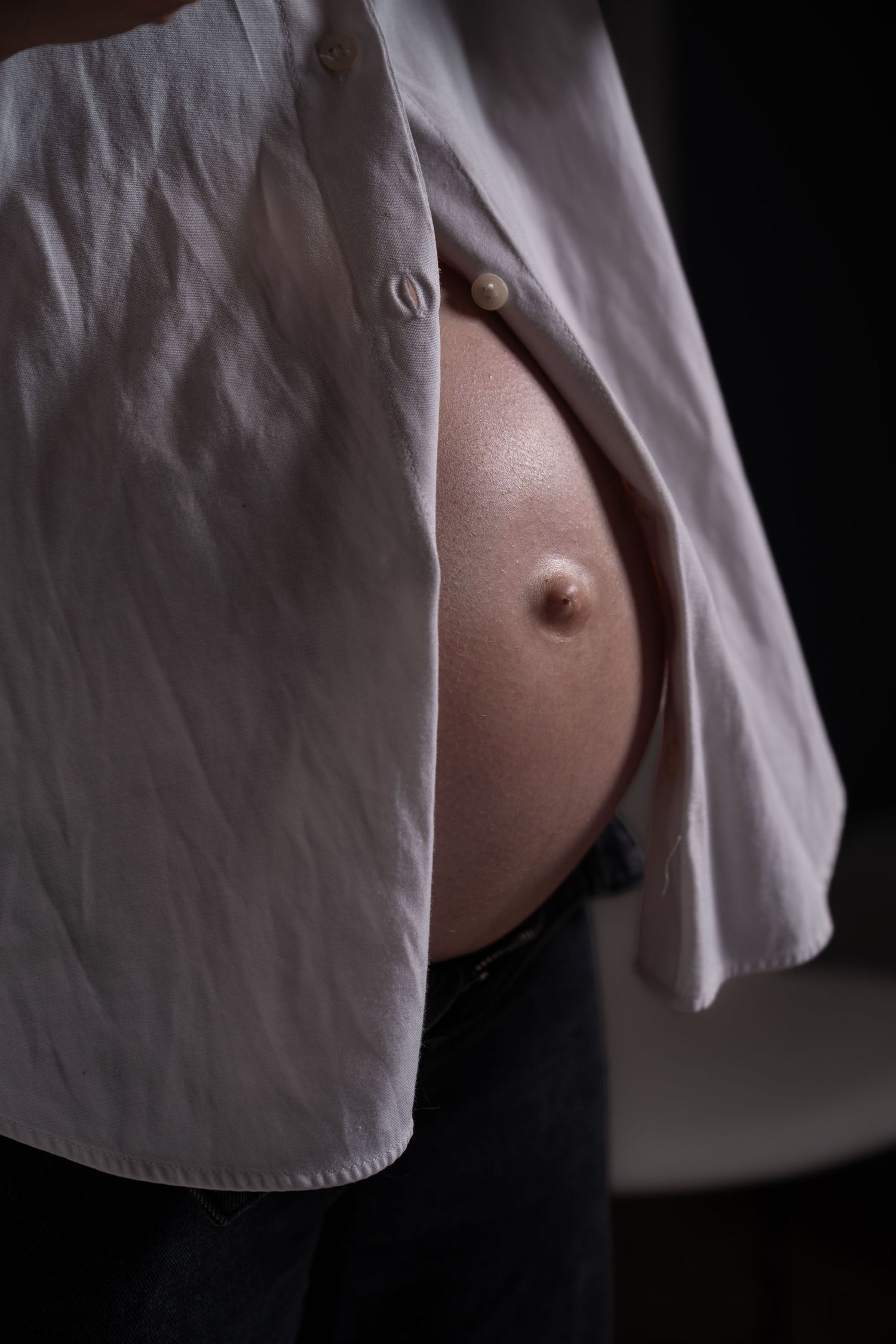 Close-up of a pregnant woman's bare belly with an open white shirt, highlighting her third trimester bump.