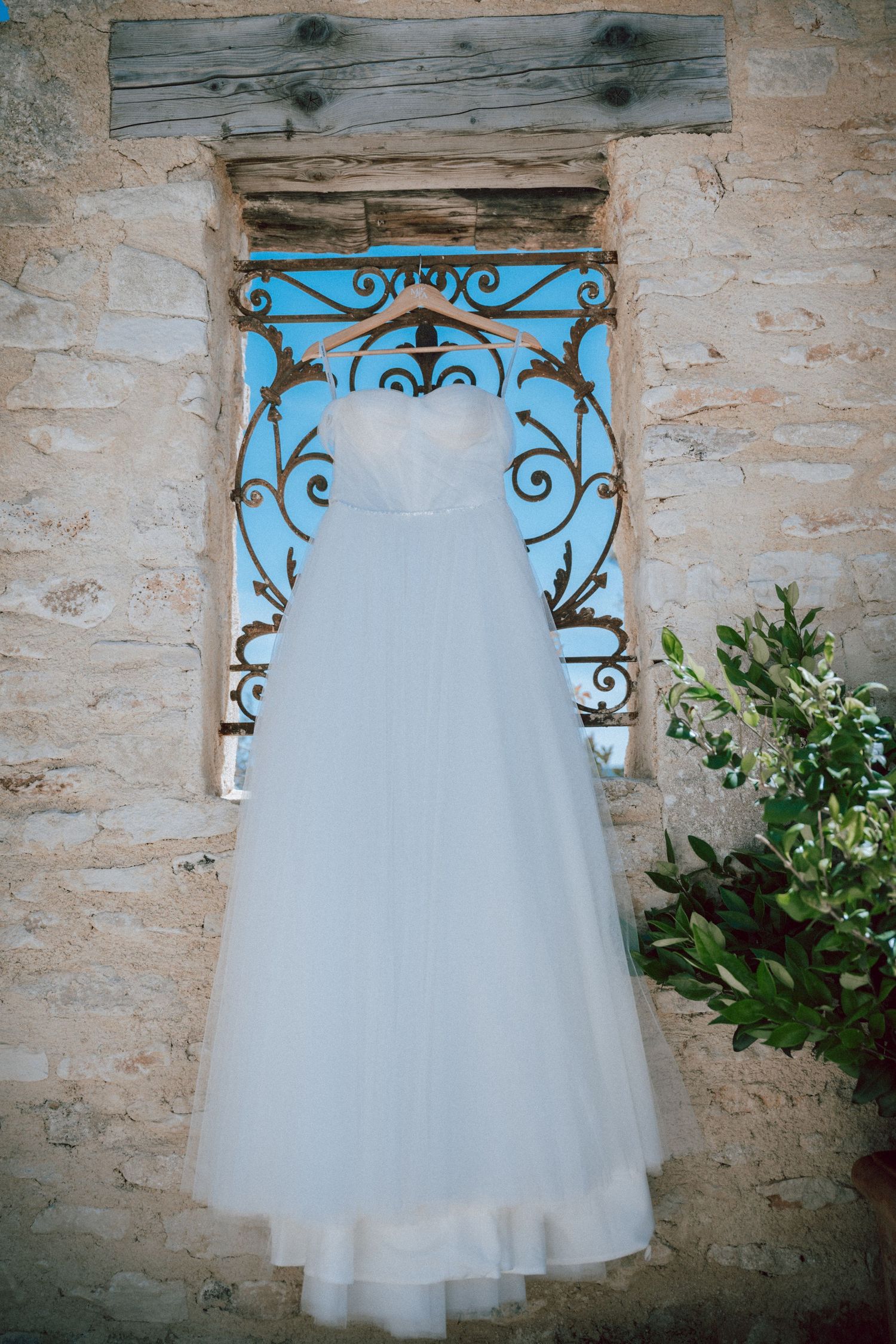 White wedding dress hanging on ornate blue iron gate against rustic stone wall.