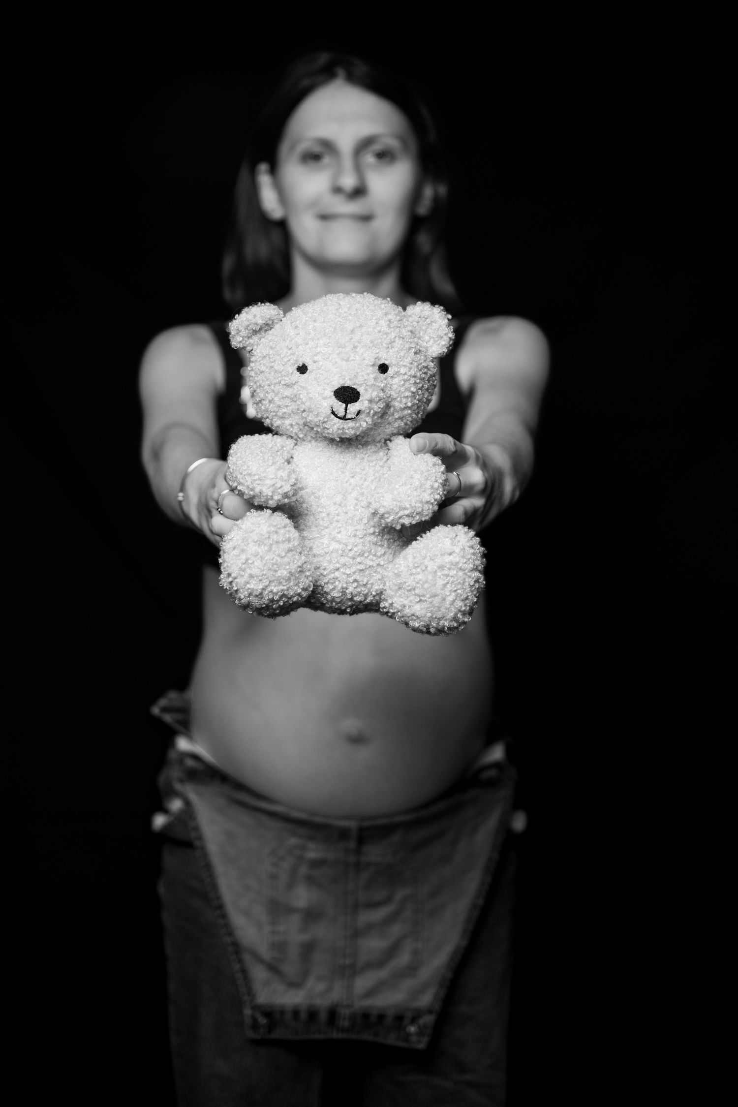 Pregnant woman holding white teddy bear toward camera in black and white maternity studio photo.