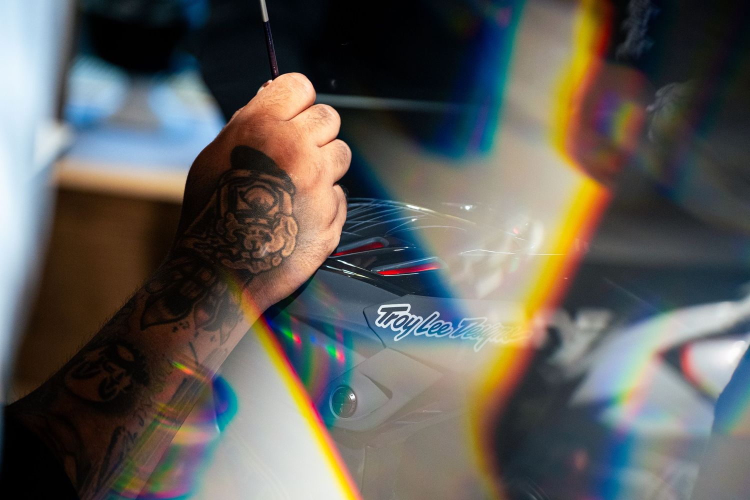 Tattooed hand painting Troy Lee Designs logo on a helmet with prismatic light effects.