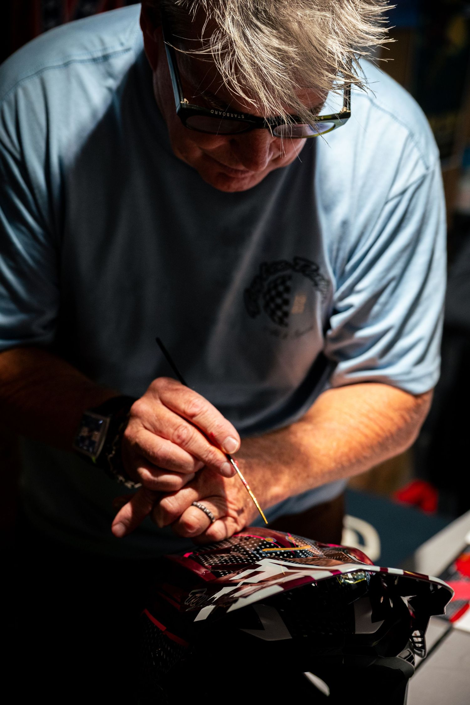 Man with sunglasses carefully painting a racing helmet with a fine brush outdoors in bright sunlight.