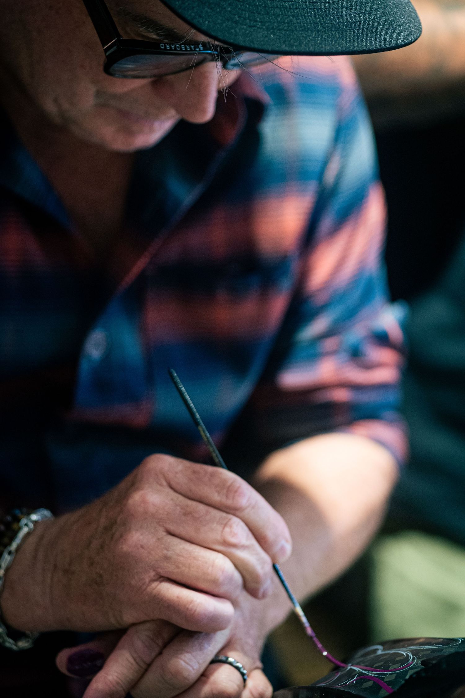 Man in plaid shirt and cap carefully painting with a fine brush, focused on detailed artwork.