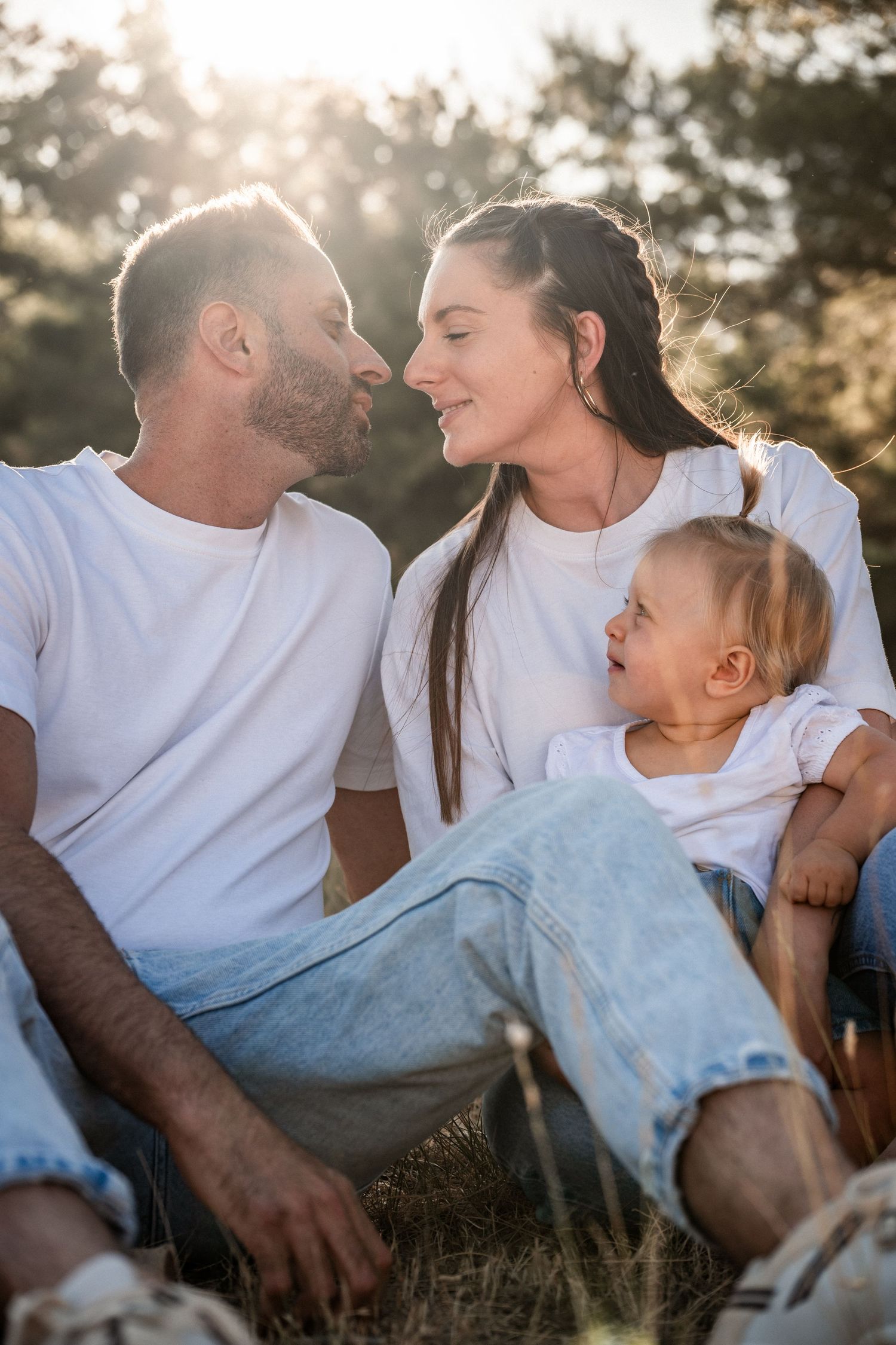 Happy family in white t-shirts sitting outdoors in golden sunlight, parents sharing a romantic moment while toddler smiles.