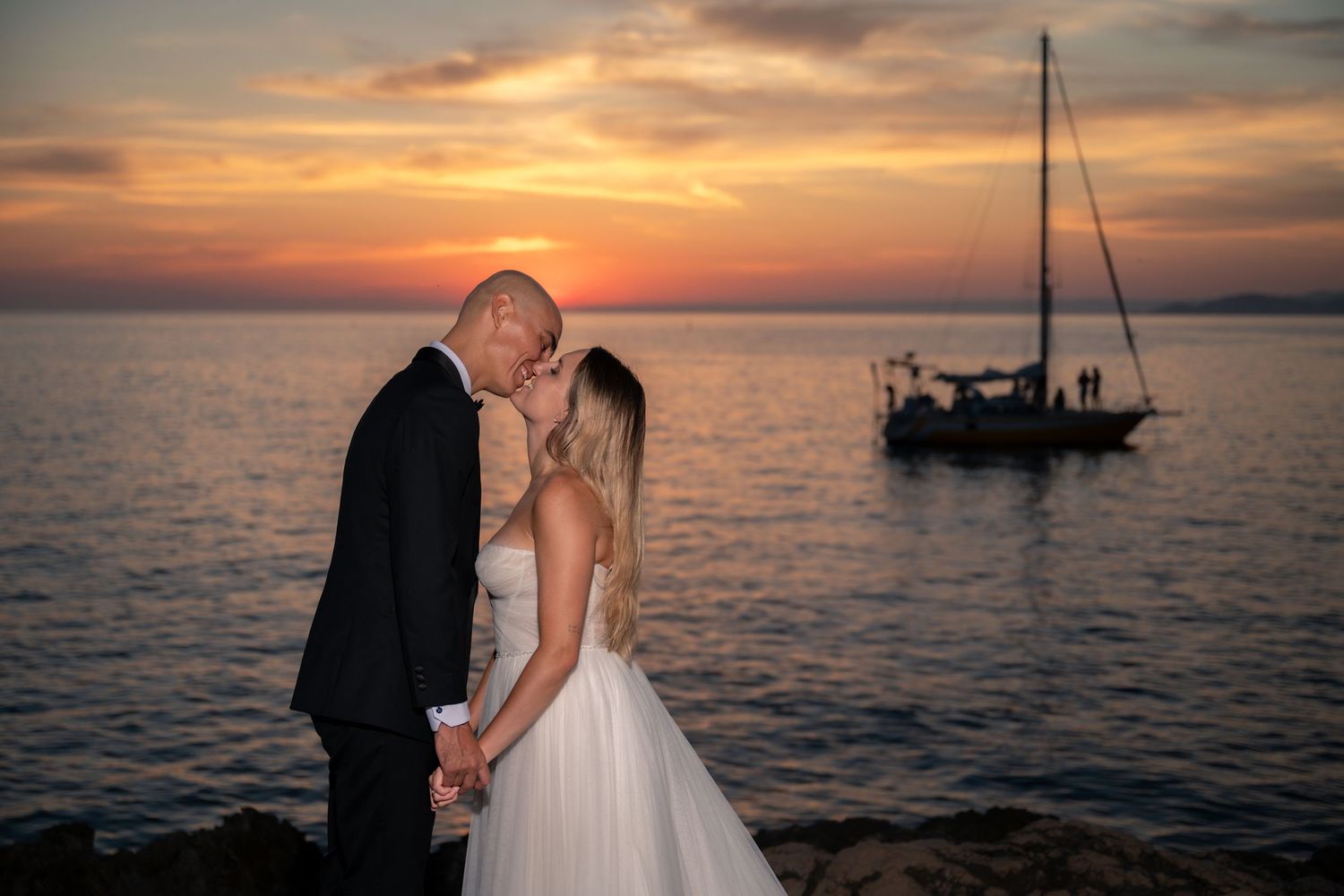 Bride and groom share a romantic kiss on rocky shore at sunset with sailboat silhouetted behind them.