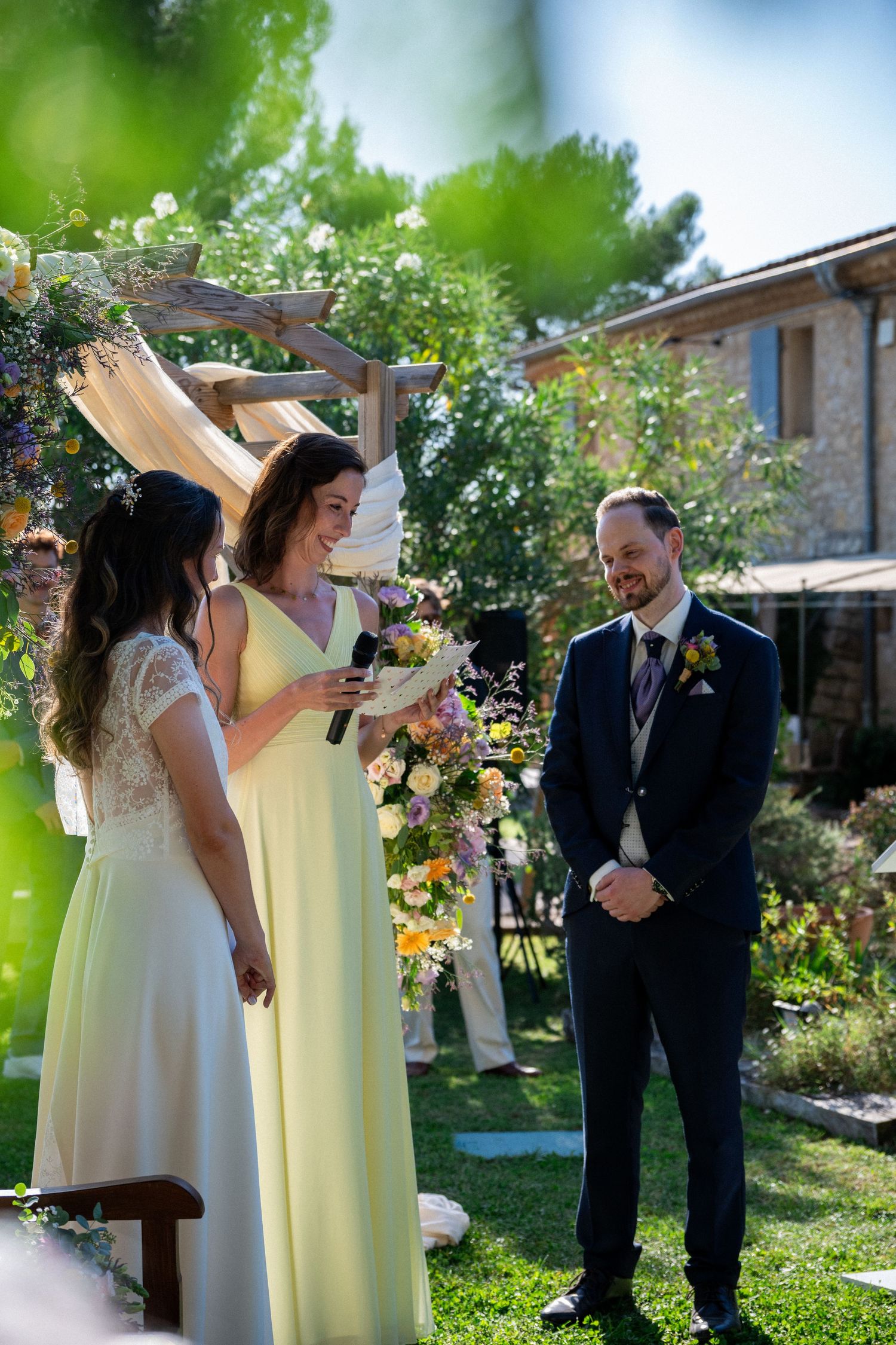 Bride reads vows to groom at outdoor wedding ceremony, flanked by bridesmaid in yellow dress under floral arch.
