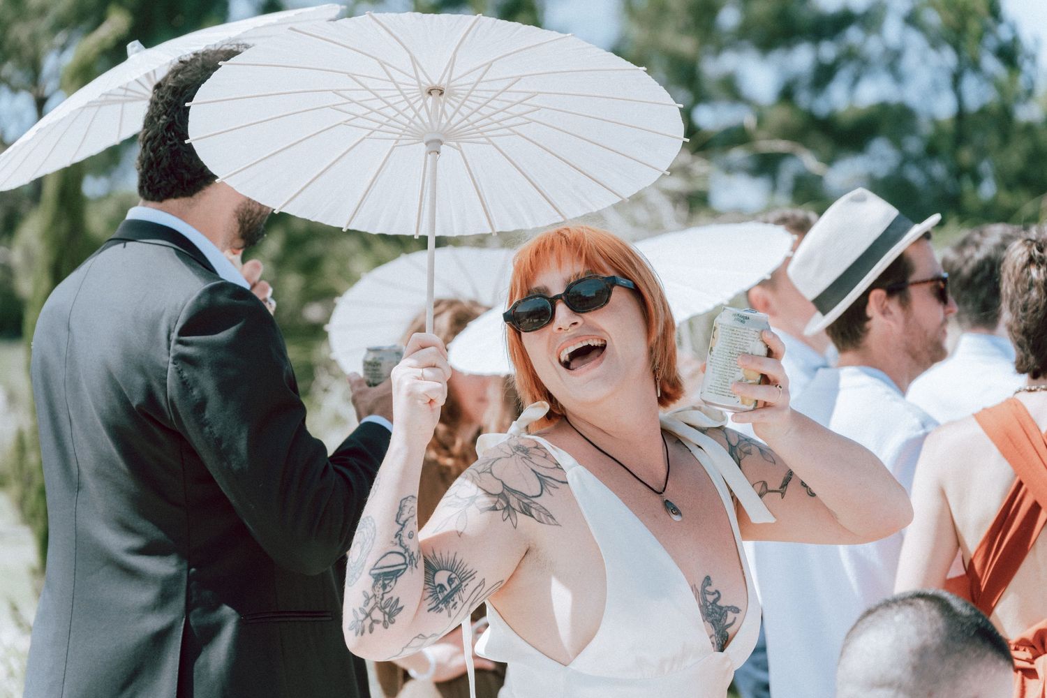 Tattooed woman laughing joyfully holding white parasol at outdoor wedding celebration.