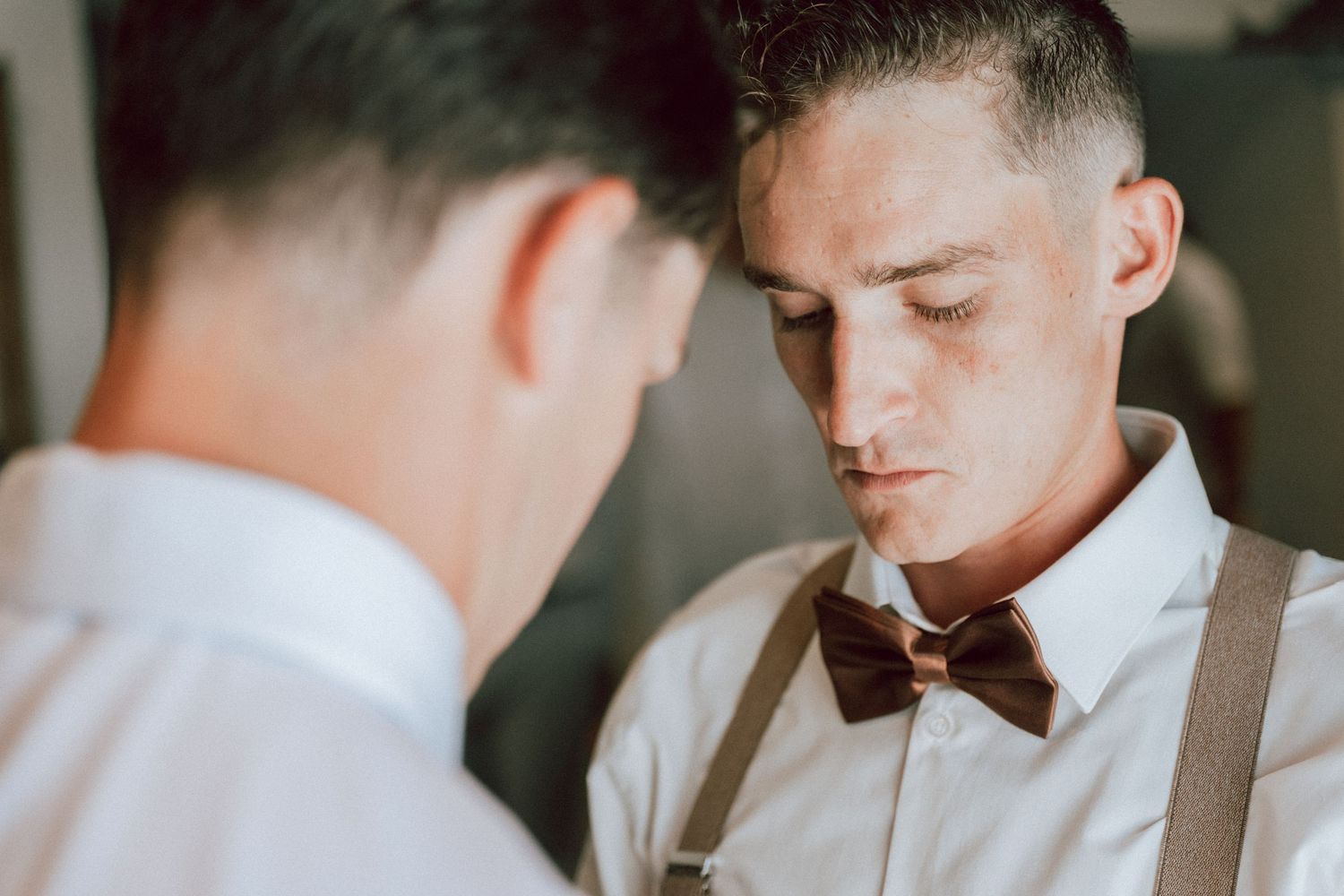 Groom adjusting bow tie with suspenders assisted by groomsman during wedding preparation.