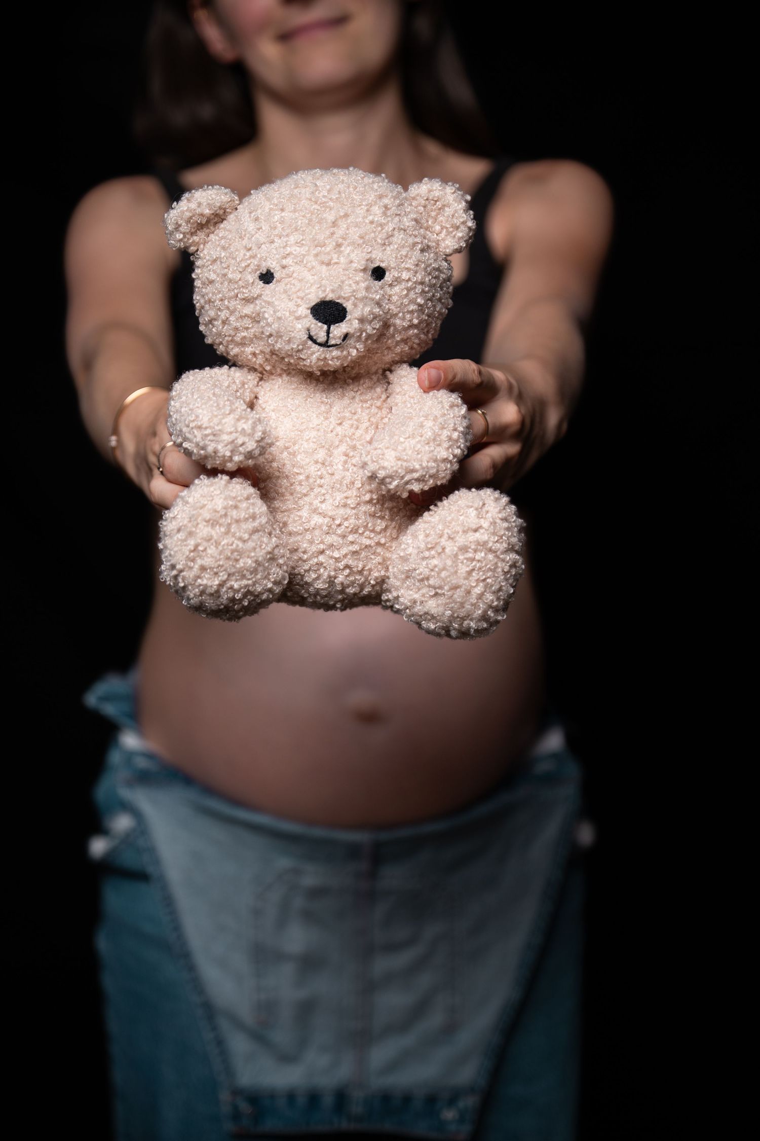 Pregnant woman in jeans holding a soft teddy bear in front of her bare baby bump.