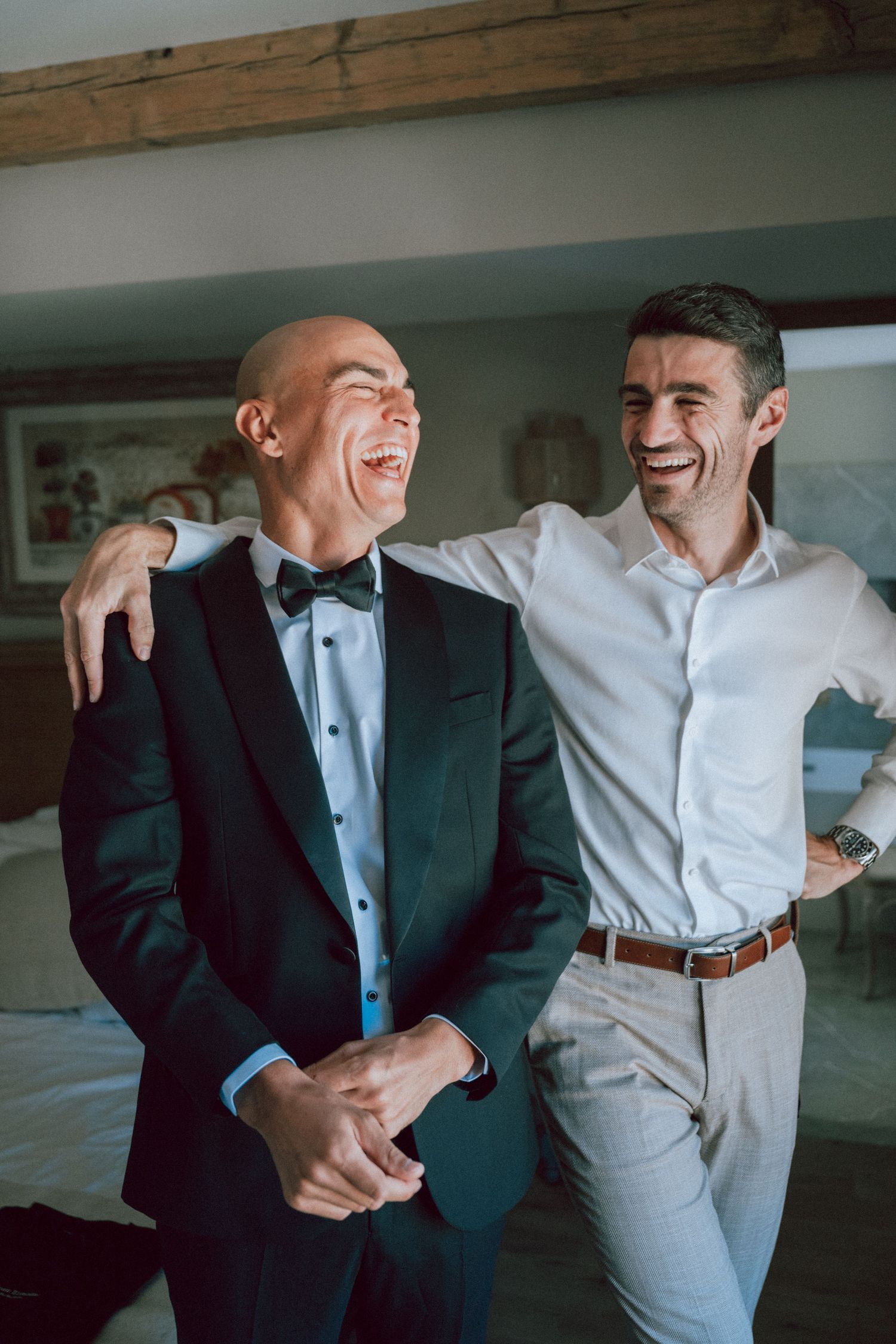 Groom in black tuxedo laughs joyfully with groomsman in white shirt during wedding prep.