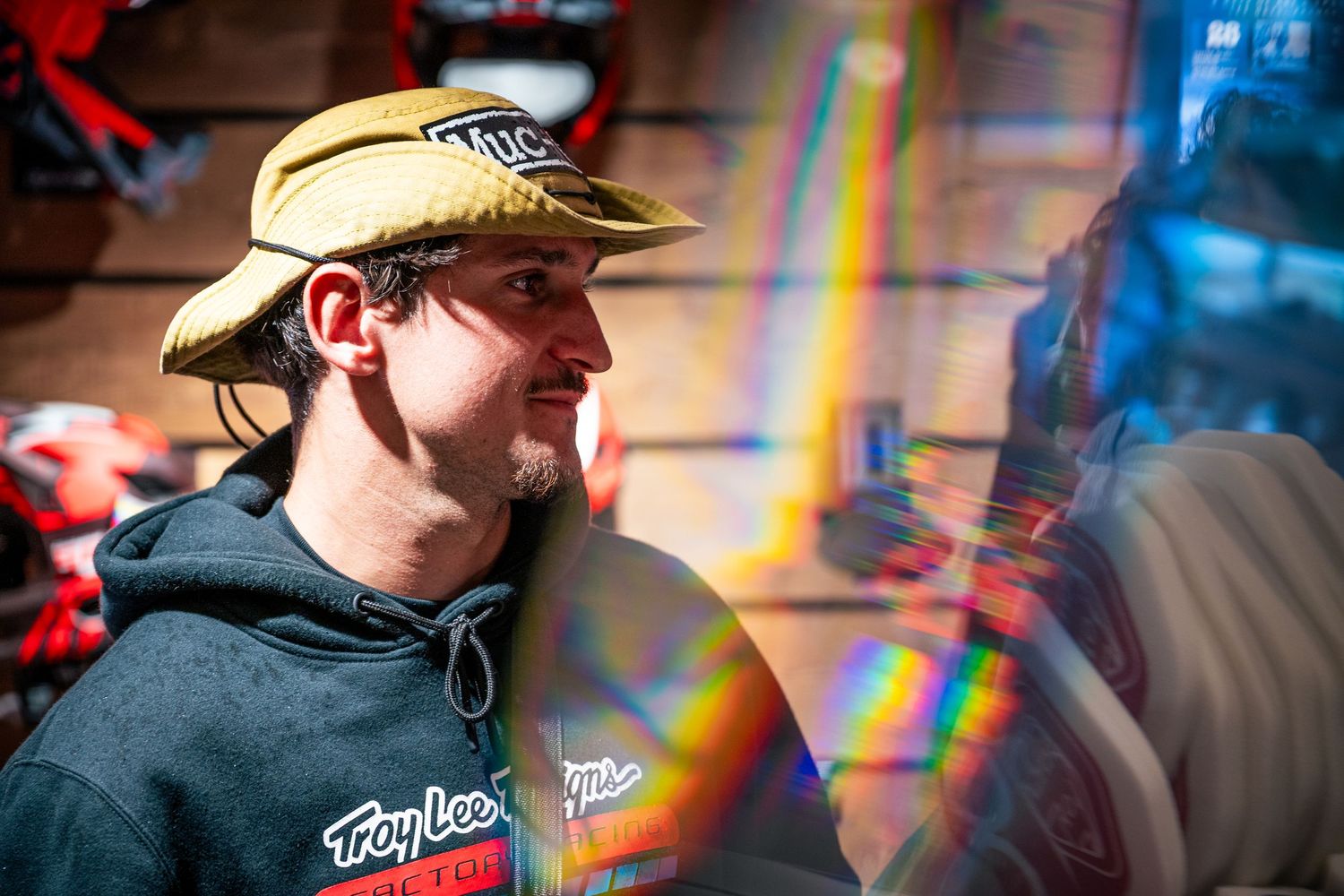 Man wearing a Troy Lee Designs hoodie and yellow bucket hat standing near a colorful rainbow light reflection.