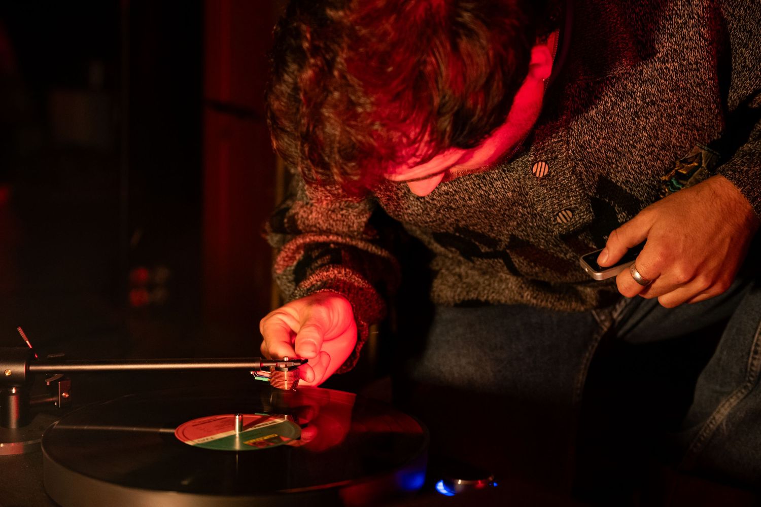 Person adjusting a vinyl record on a turntable under red ambient lighting at a music event.