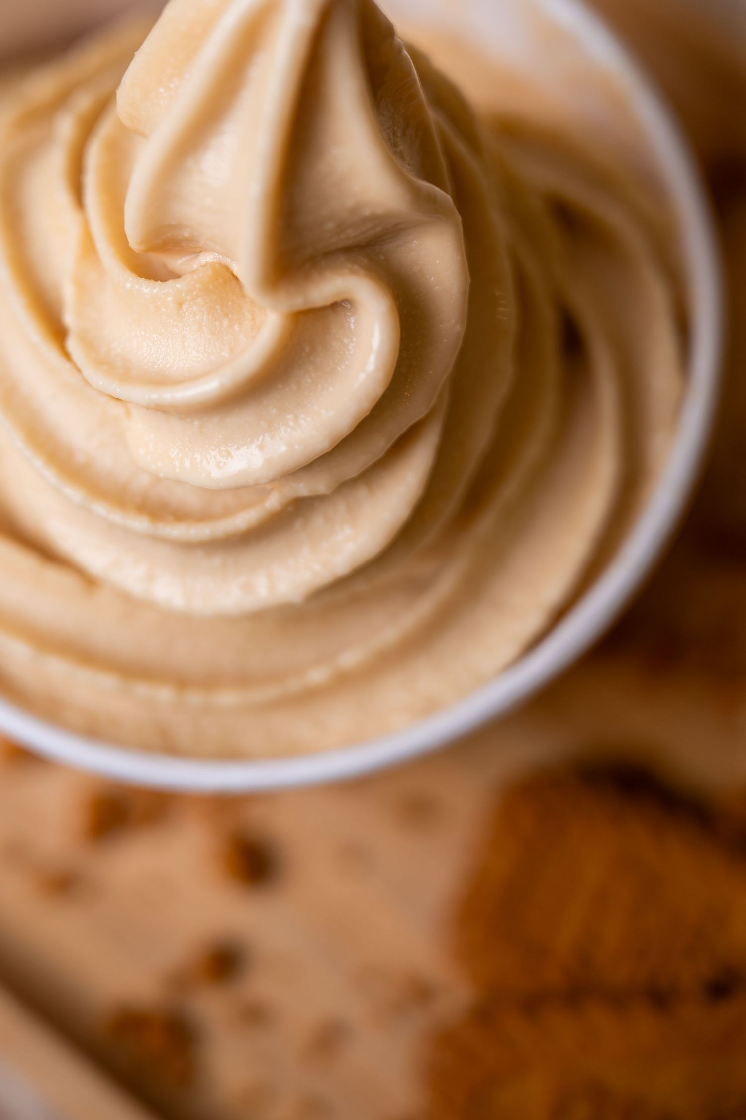 Close-up of a swirled soft serve ice cream in a white cup with cookies in the background.