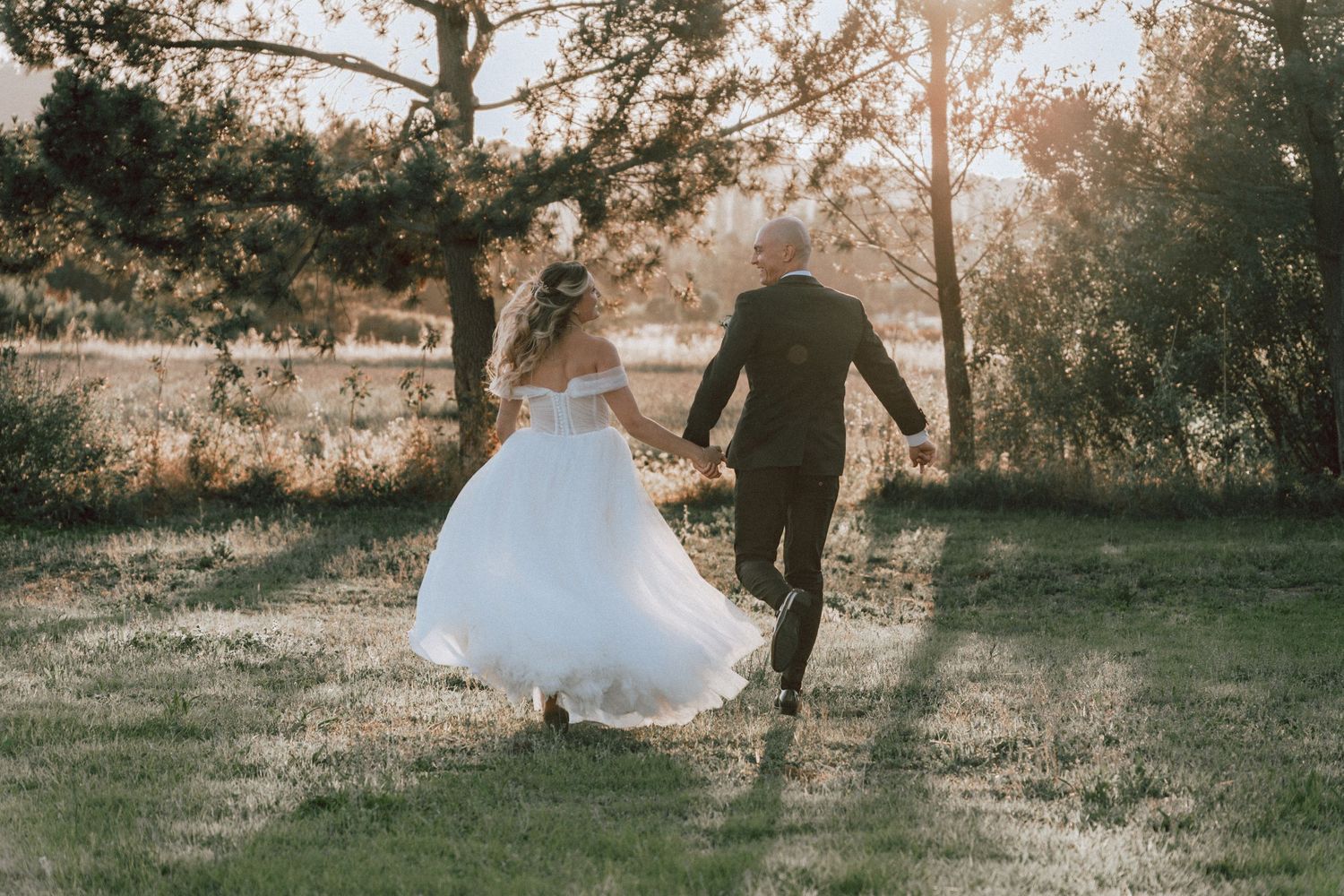 Newlywed couple runs hand-in-hand through sunlit field, bride in white gown at golden hour.