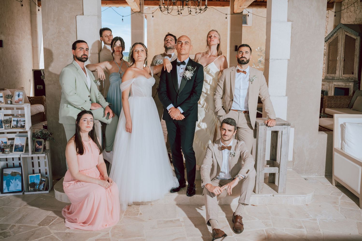 Wedding party group photo with bride in white gown and groom in black tuxedo surrounded by bridesmaids and groomsmen indoors.