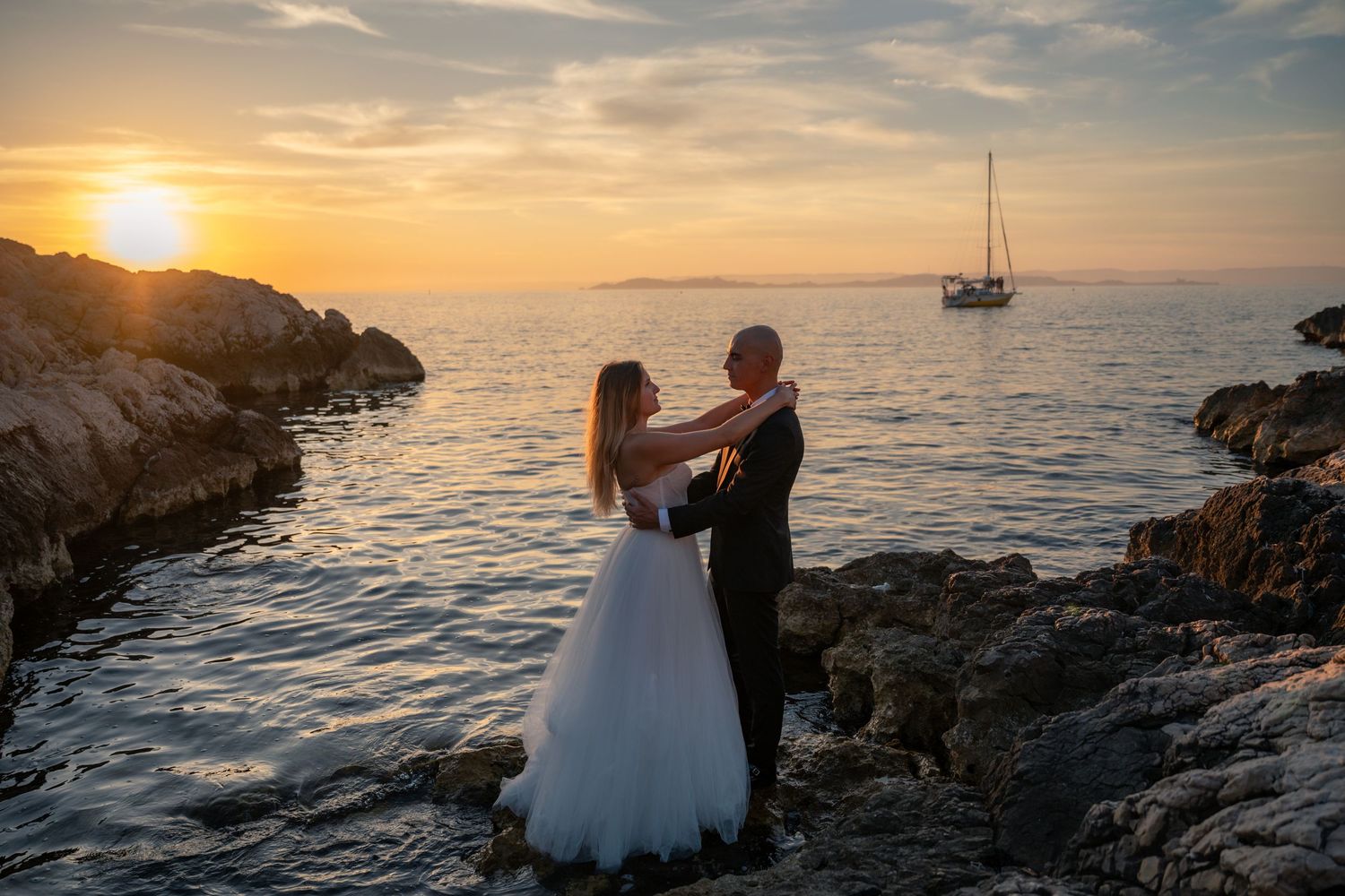 Bride and groom dancing on rocky shore with sailboat and glowing sunset.