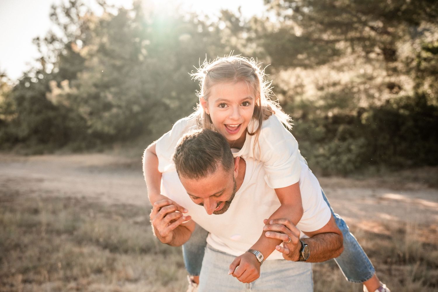 Happy couple playing outdoors in a sunny field, woman piggyback riding on mans back, smiling joyfully.
