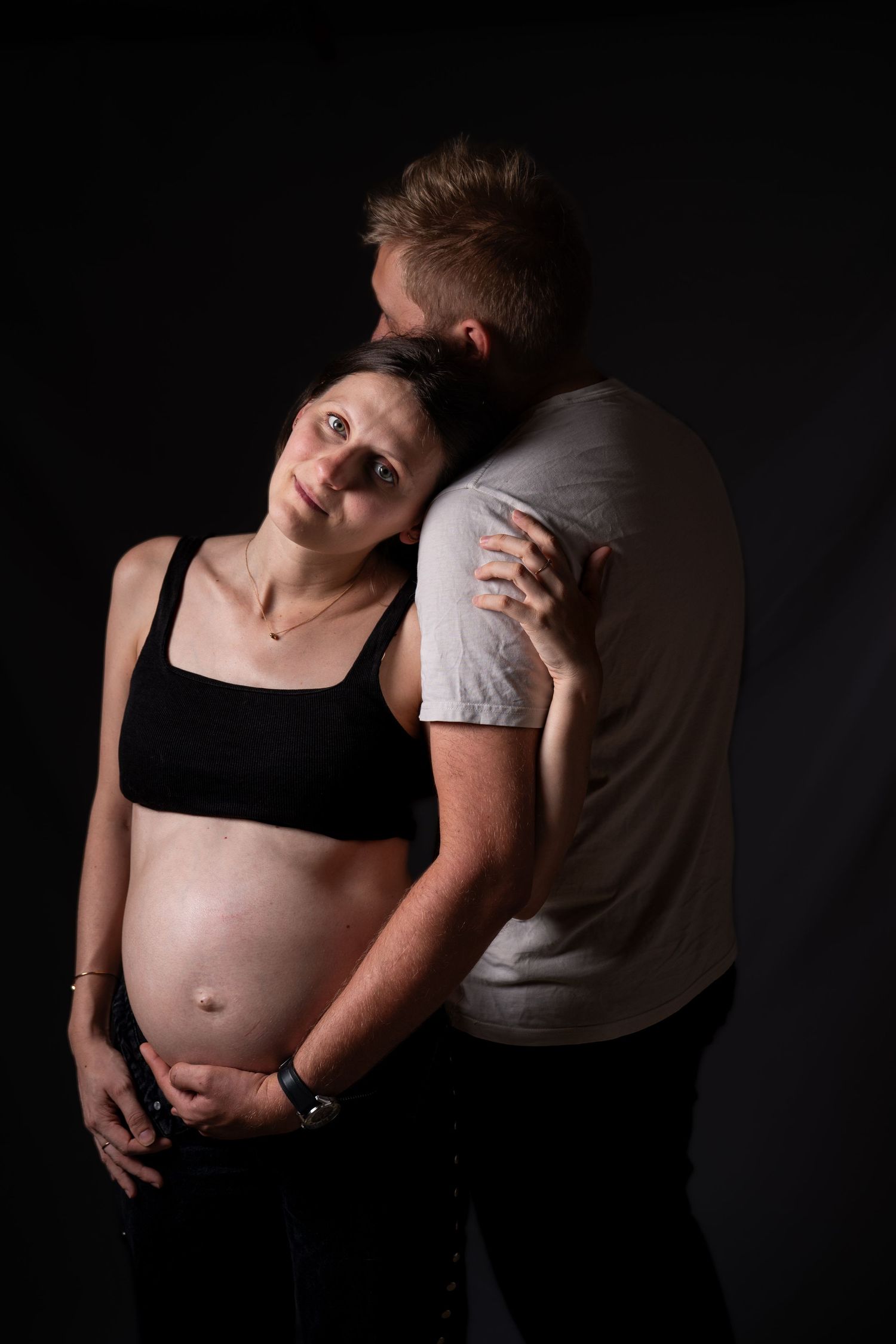 Pregnant woman in black crop top leaning against her partners chest in a dark studio portrait setting.