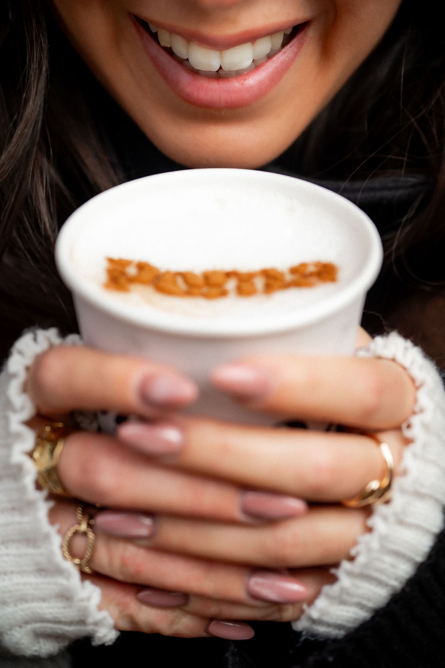 Smiling woman holding a frothy cappuccino cup with decorative text latte art design.