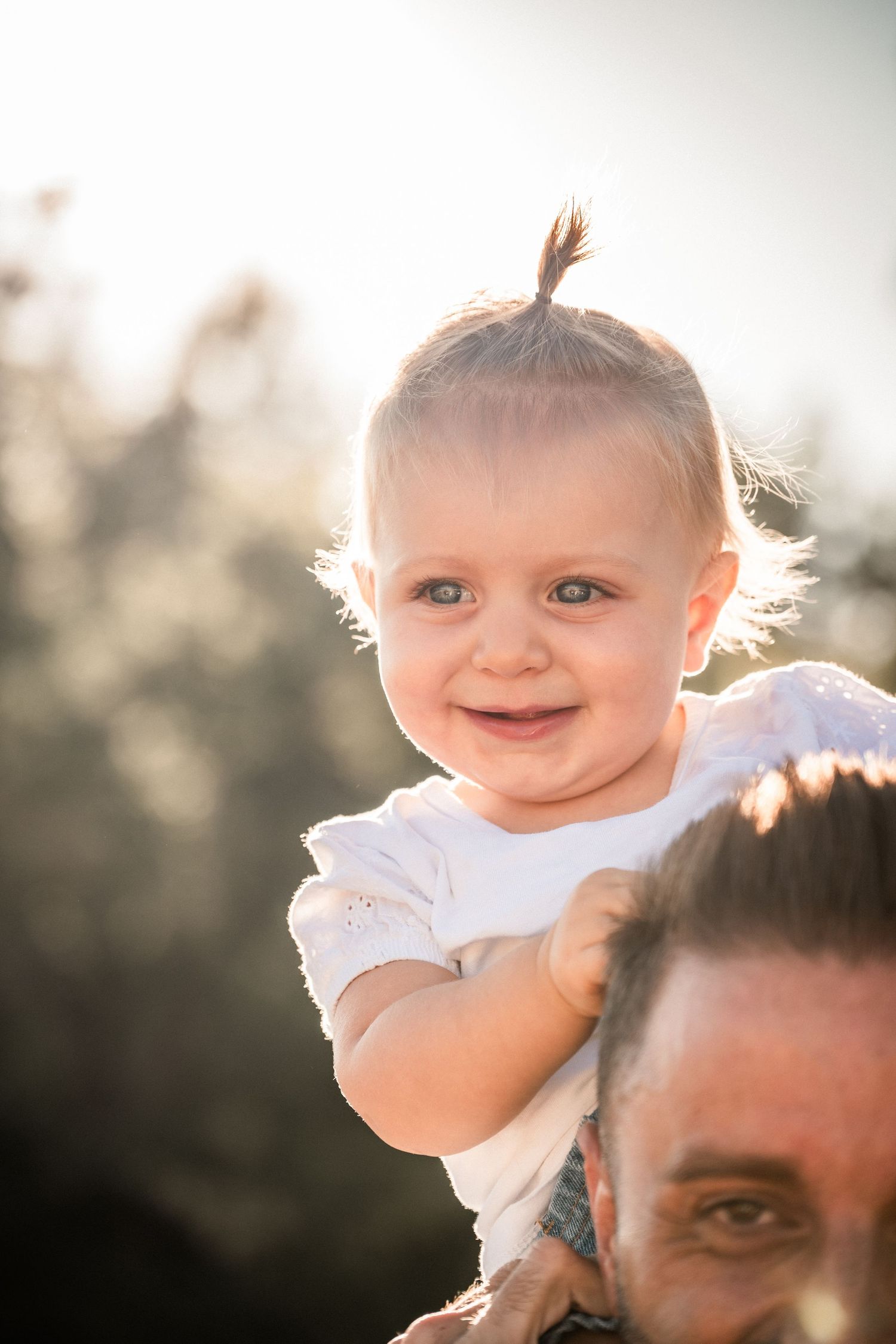 Smiling baby with tiny ponytail perched on fathers shoulders in sunny outdoor setting.